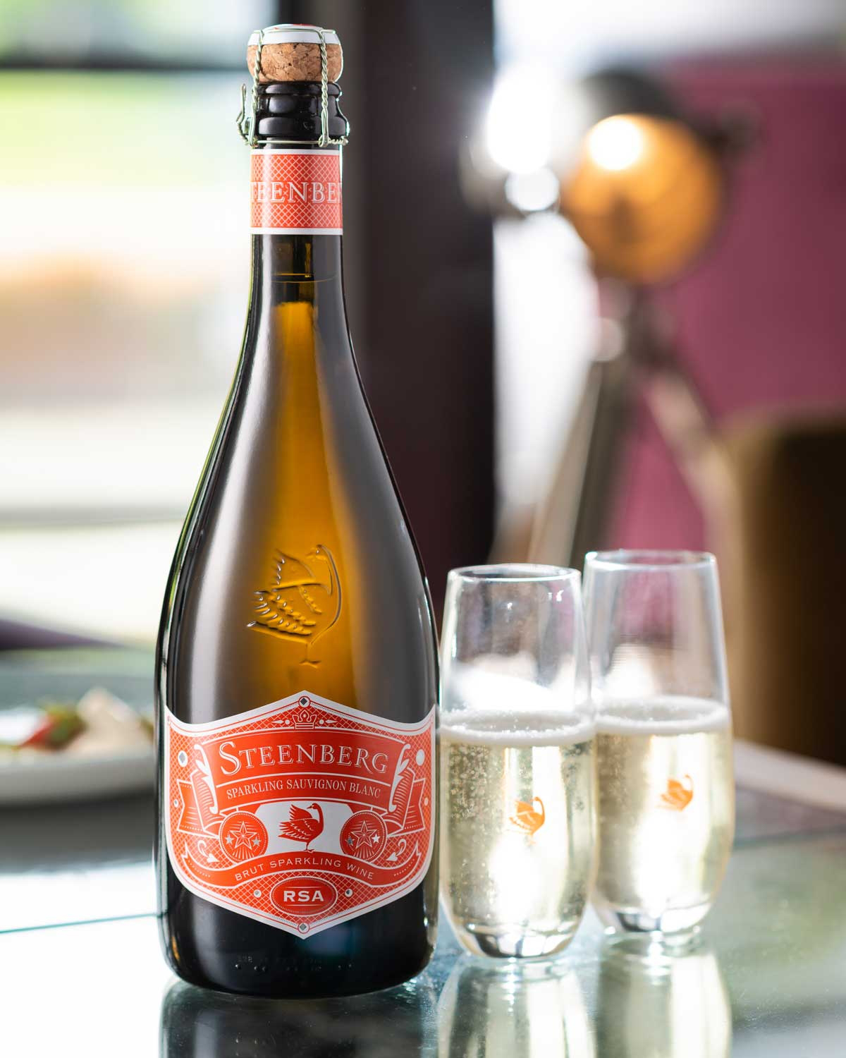 Eine Flasche Steenberg Sparkling Sauvignon Blanc mit einem orange-weißen Etikett, einem mit Draht gesicherten Korken und einem geätzten Vogelmotiv auf dem Glas. Das Etikett weist darauf hin, dass es sich um einen Brut-Schaumwein aus Südafrika handelt.