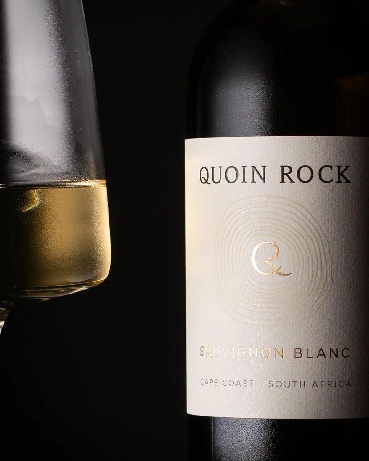 Eine Flasche Quoin Rock Sauvignon Blanc mit Goldfolienverschluss und einem cremefarbenen Etikett, auf dem das Logo, der Name des Weins und die Herkunft angegeben sind: Cape Agulhas, Südafrika.