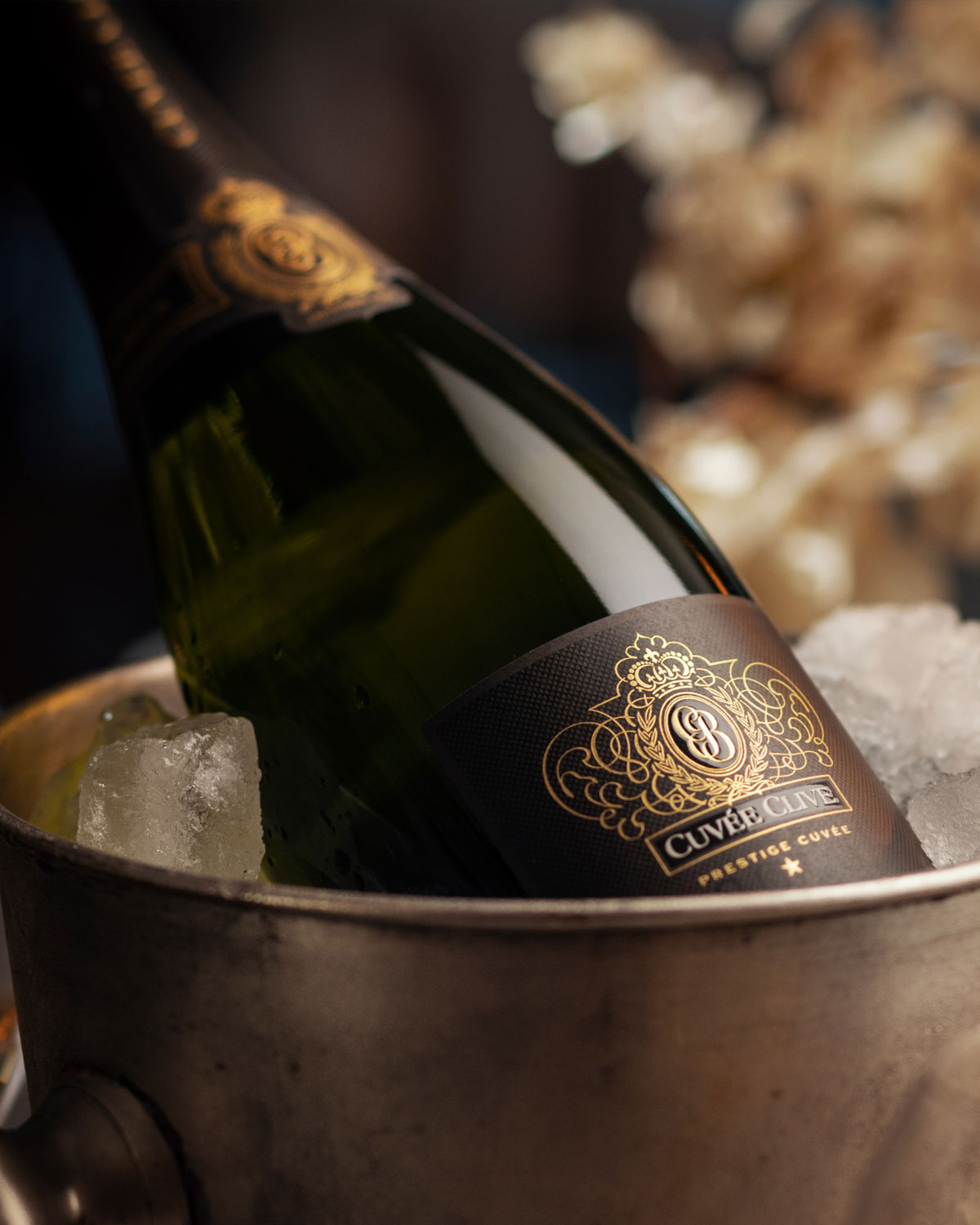 Eine Sektflasche mit der Aufschrift Cuvée Cave ruht in einem metallenen Eiskübel, umgeben von Eiswürfeln, mit weicher, warmer Beleuchtung und verschwommenen Blumen im Hintergrund.