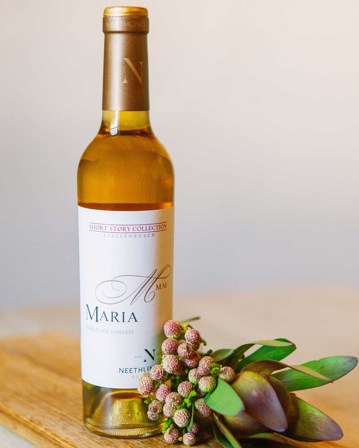 Eine Flasche Neethlingshof Maria Noble Spätlese steht neben der dazu passenden cremefarbenen zylindrischen Schachtel mit goldener und hellbrauner Beschriftung und dekorativen Mustern.