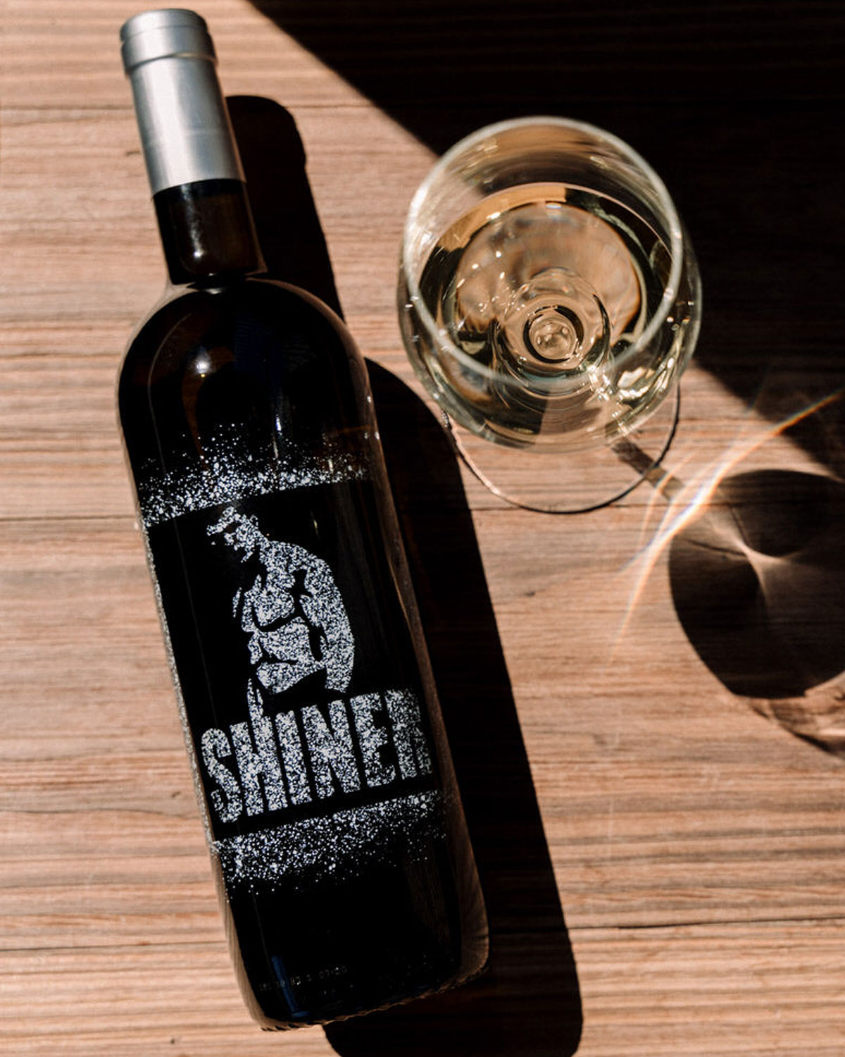 Eine Klarglasflasche mit einem silbernen Deckel, auf dessen Etikett ein sich beugender Bodybuilder und das Wort SHINER in fetten, funkelnden Buchstaben abgebildet sind. Die Flasche scheint ein helles Getränk zu enthalten.