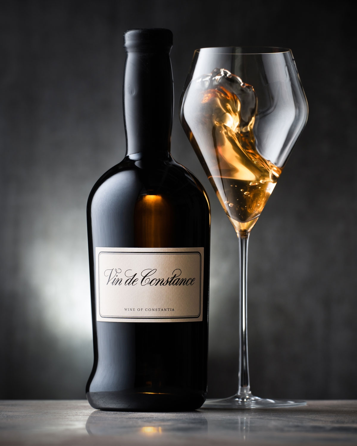 Eine Flasche Vin de Constance steht neben einem Glas, in dem goldener Wein schwappt, vor einem dunklen, sanft beleuchteten Hintergrund.