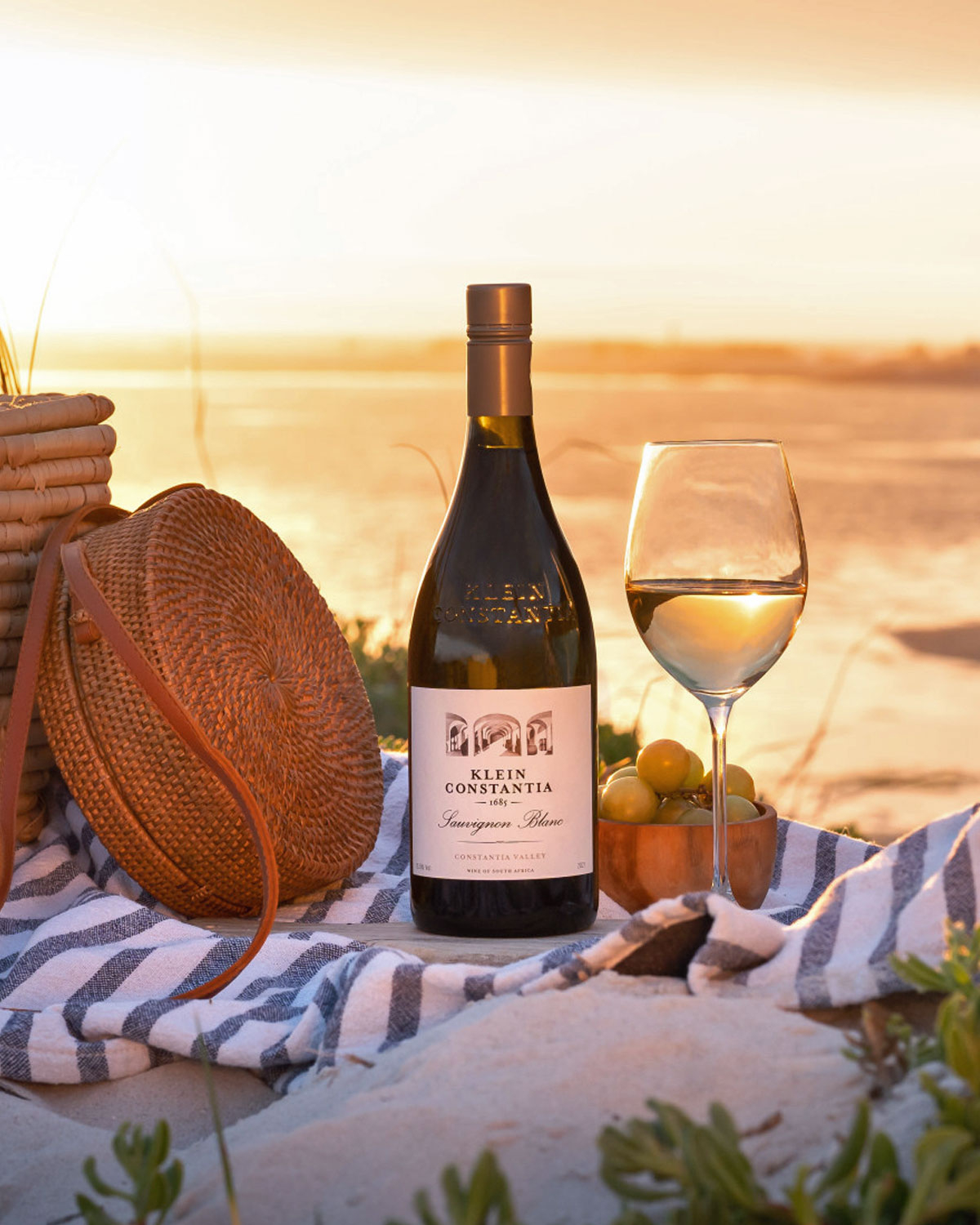Eine Flasche Klein Constantia Sauvignon Blanc, ein Glas Weißwein, eine Schale mit grünen Weintrauben, eine geflochtene Tasche und ein gestreiftes Handtuch stehen bei Sonnenuntergang auf einem Felsen mit Blick auf ein Gewässer.