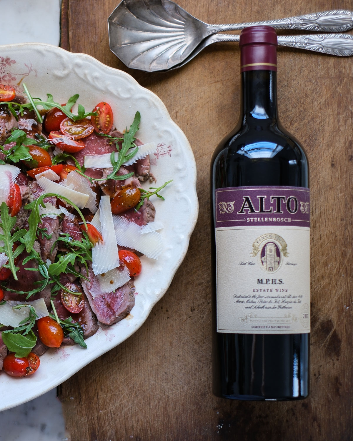 Eine Flasche Alto Stellenbosch Rotwein steht auf einer Holzplatte neben einem Blumenteller mit einem frischen Salat aus Rucola, Kirschtomaten, gehobeltem Käse und Rindfleischscheiben.