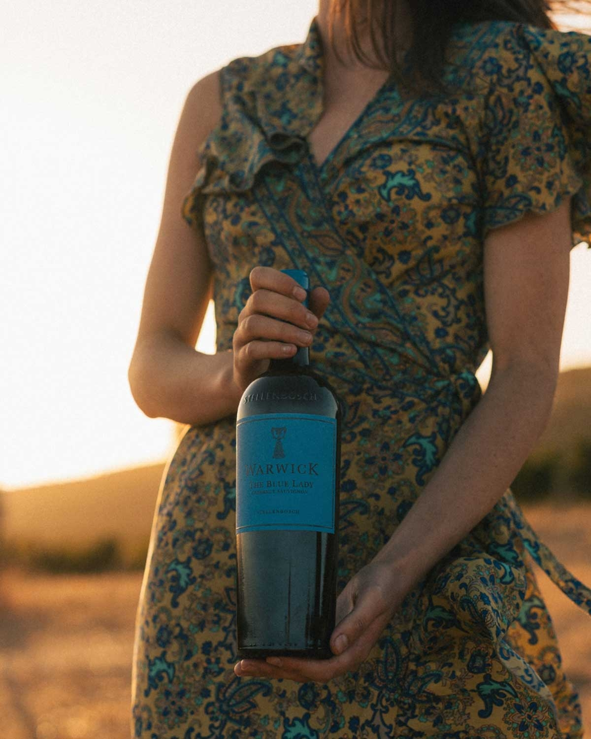 Eine Person in einem geblümten Kleid steht bei Sonnenuntergang im Freien und hält eine Flasche Warwick-Wein mit einem blauen Etikett in beiden Händen. Das Gesicht der Person ist nicht zu erkennen.