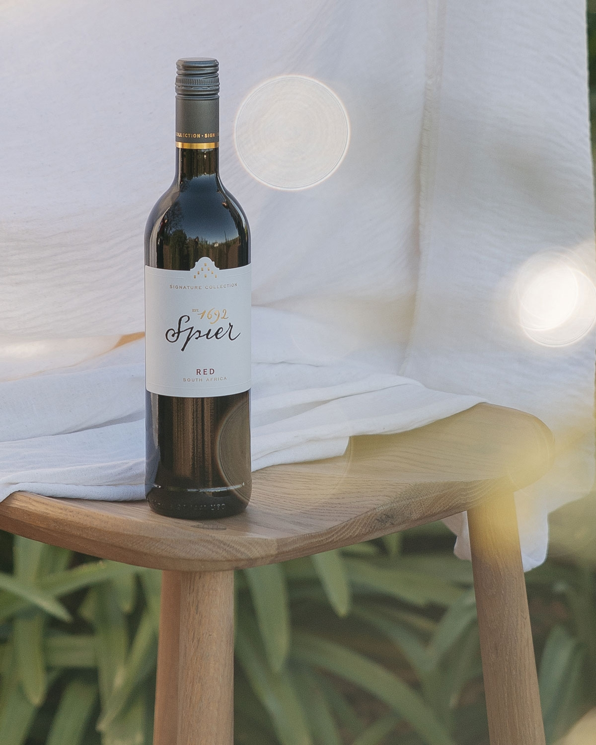 Eine Flasche Spier 1692 Rotwein steht auf einem Holzhocker, der mit einem weißen Tuch bedeckt ist. Im unscharfen Hintergrund sind Grünpflanzen zu sehen, und das weiche Sonnenlicht erzeugt sanfte Lichtreflexe.