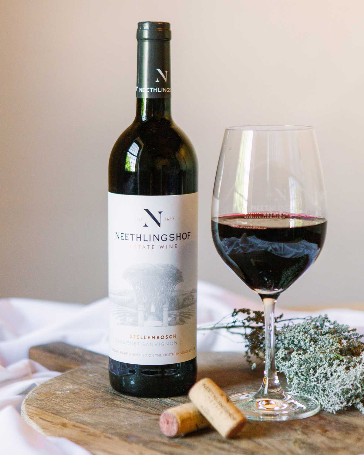 Eine Flasche Neethlingshof Estate Wine, Stellenbosch Cabernet Sauvignon, mit einem beigen Etikett mit einer Zeichnung von Bäumen und Gebäuden des Weinguts und Text in Schwarz und Gold.