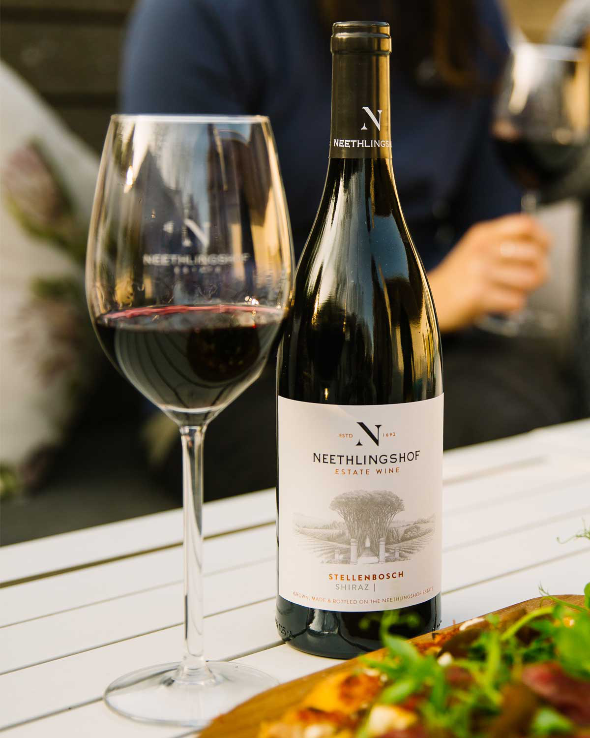 Eine Flasche Neethlingshof Estate Wine Shiraz aus Stellenbosch mit einem minimalistischen, cremefarbenen Etikett mit einer Illustration des Weinbergs und dem Namen und Emblem des Winzers in eleganten Buchstaben.