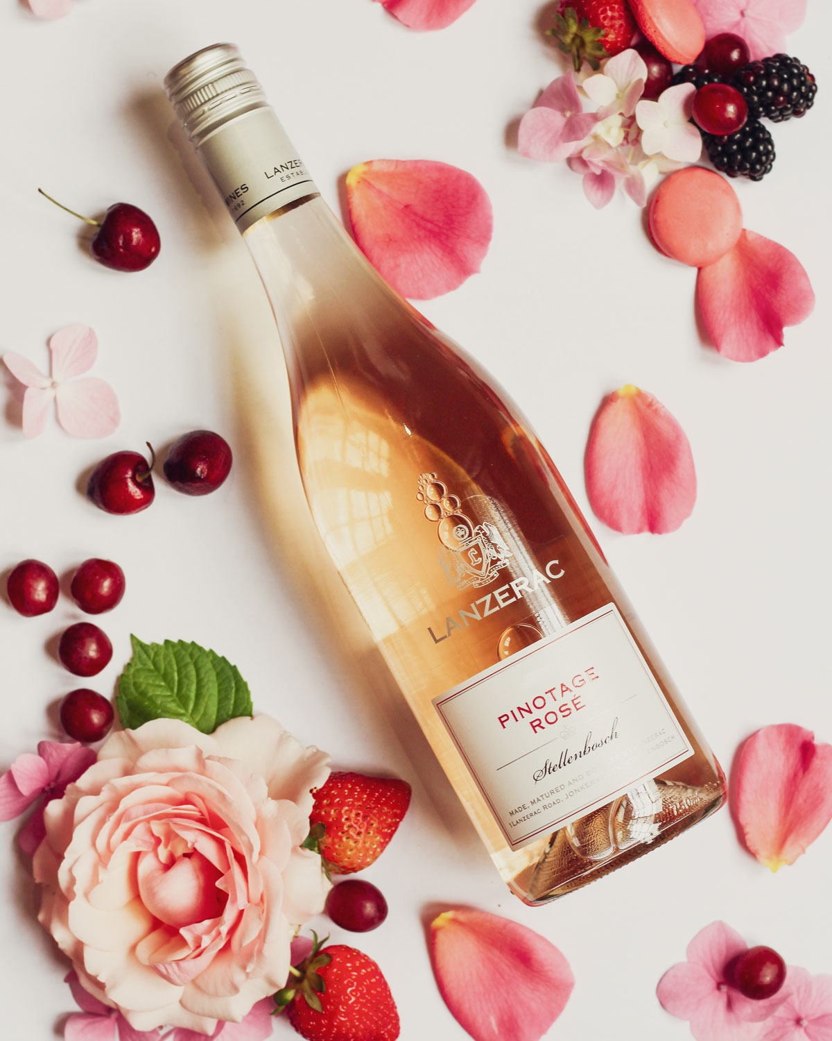 Eine Flasche Lanzerac Pinotage Rosé liegt auf einer weißen Fläche, umgeben von rosa Rosenblättern, frischen Erdbeeren, Kirschen, Himbeeren und einer großen rosa Rosenblüte.