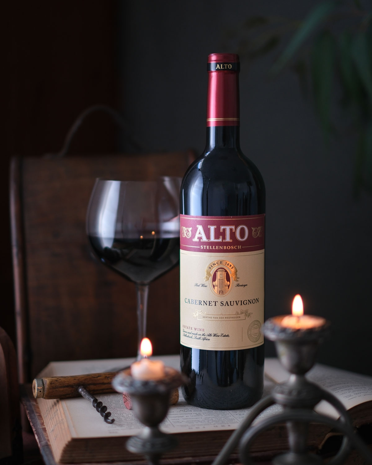 Eine Flasche Alto Cabernet Sauvignon steht auf einem aufgeschlagenen Buch neben einem gefüllten Weinglas, zwei brennenden Kerzen und einer hölzernen Pfeife und schafft eine gemütliche, alte Atmosphäre.