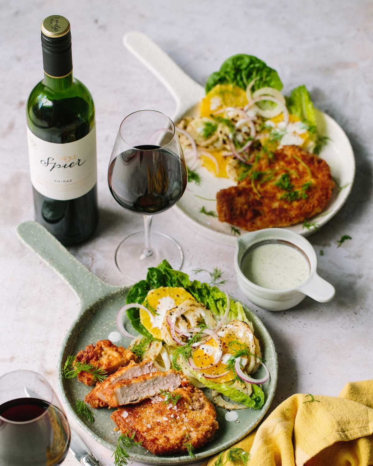 Zwei Teller mit knusprig panierten Schnitzeln, grüner Salat mit Orangenscheiben und Zwiebeln, ein Glas und eine Flasche Rotwein, ein Krug mit cremigem Dressing und eine gelbe Serviette auf einer hellen Unterlage.