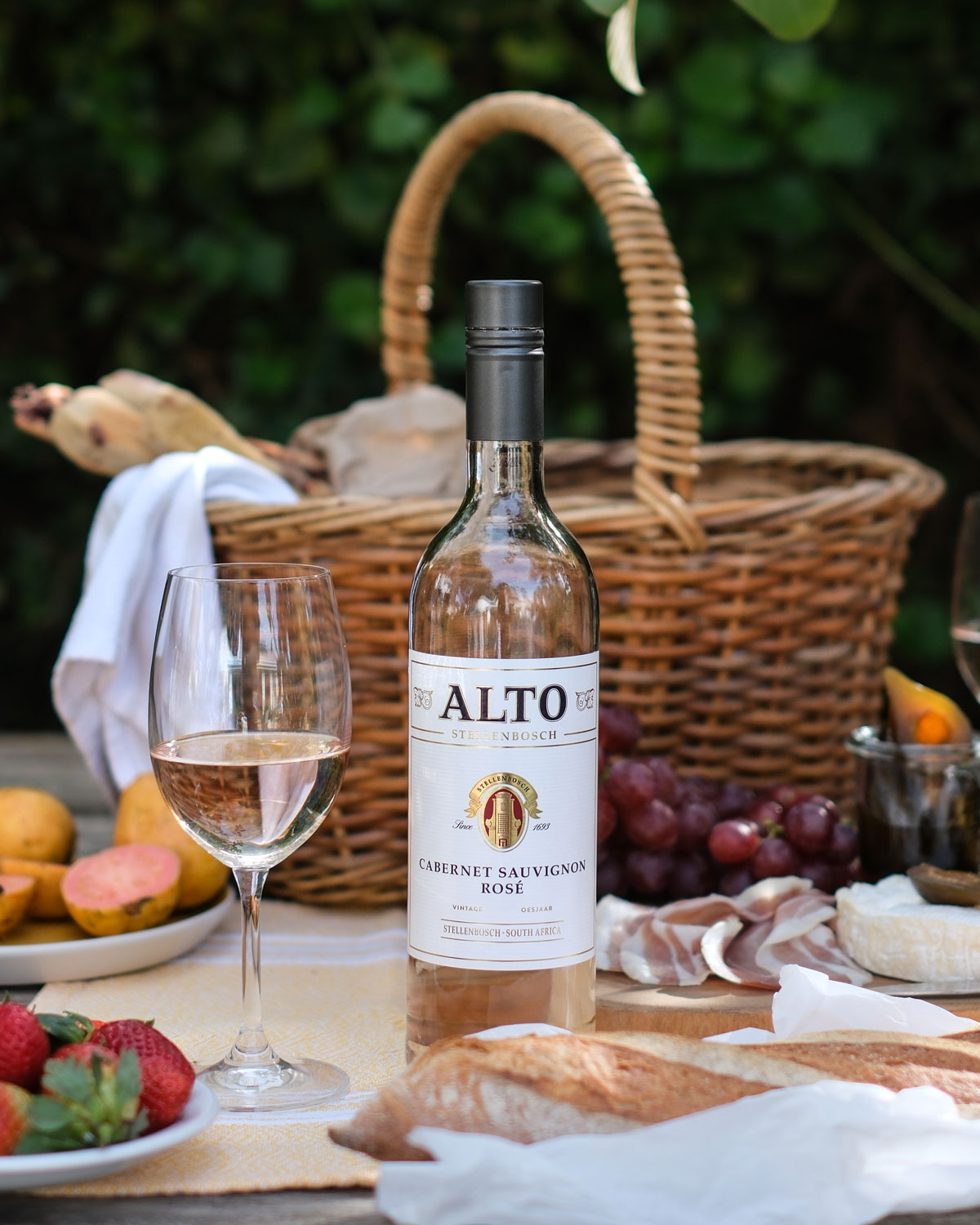 Eine Flasche Alto Cabernet Sauvignon Rosé steht neben einem gefüllten Weinglas auf einem Picknicktisch mit Brot, Obst und Fleisch. Im Hintergrund stehen ein Picknickkorb und Grünzeug.