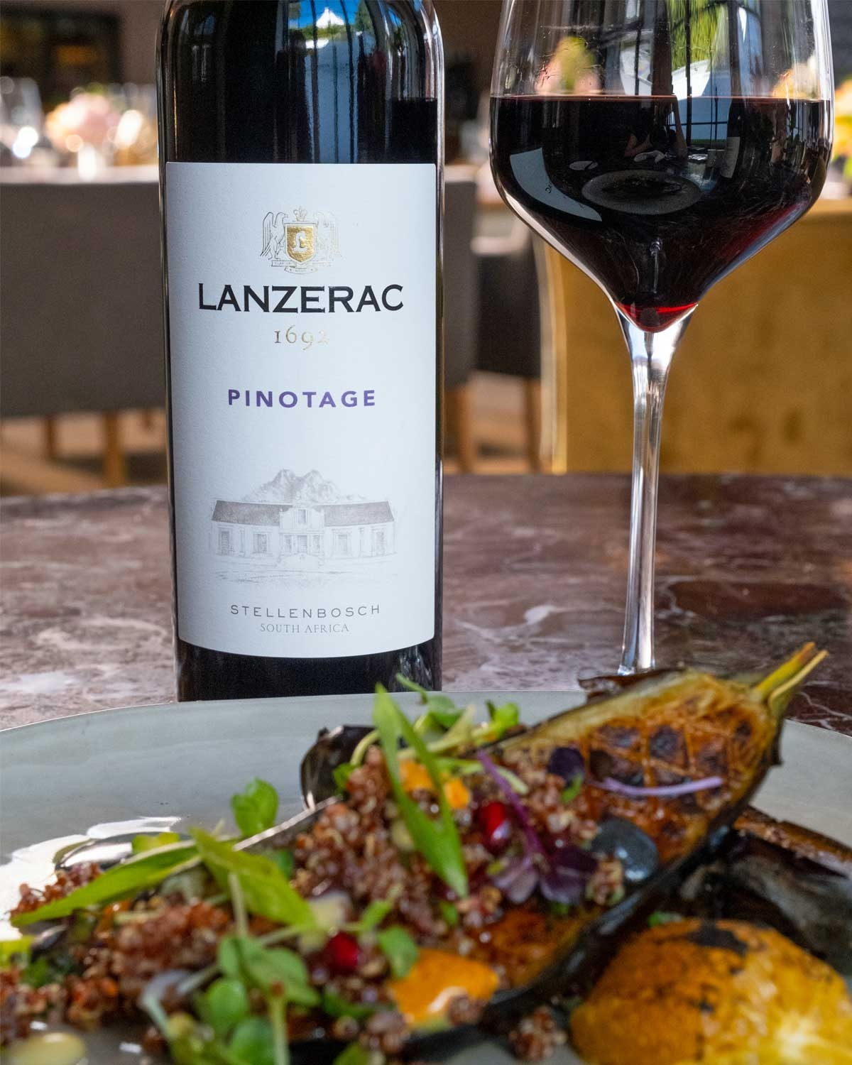 Eine Flasche Lanzerac 1692 Pinotage mit violettem Foliendach und weißem Etikett mit goldenen Akzenten, dem Lanzerac-Wappen und einer Abbildung eines Gebäudes. Der Wein stammt aus Stellenbosch, Südafrika.