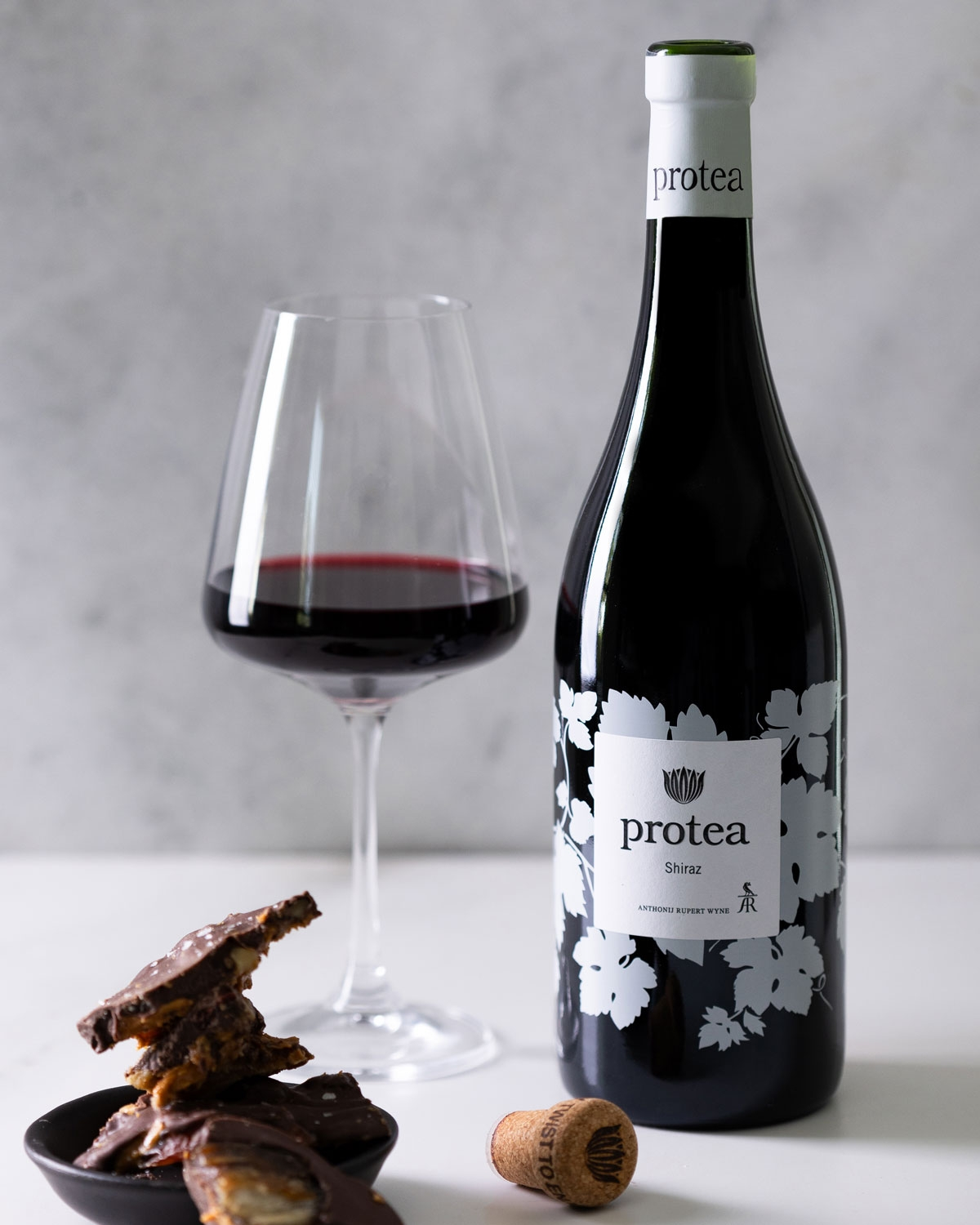 Eine Flasche Protea Shiraz Wein steht neben einem gefüllten Weinglas, einem Korken und einer kleinen schwarzen Schale mit schokoladenüberzogenen Leckereien auf einer weißen Fläche mit hellgrauem Hintergrund.