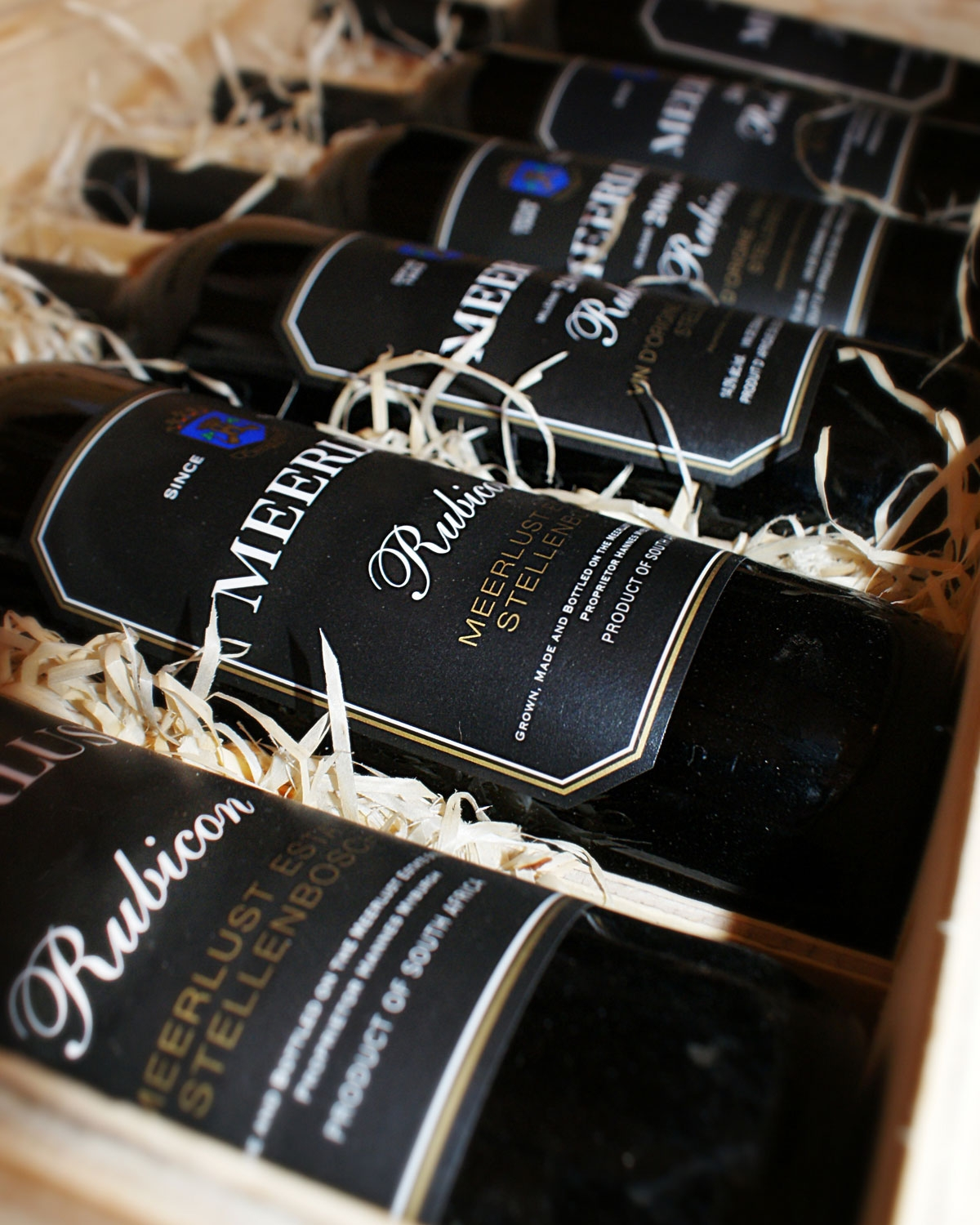 Nahaufnahme von mehreren Flaschen Meerlust Rubicon-Wein in einer Holzkiste mit Strohverpackung. Auf den schwarzen Weinetiketten steht Meerlust Rubicon Stellenbosch.