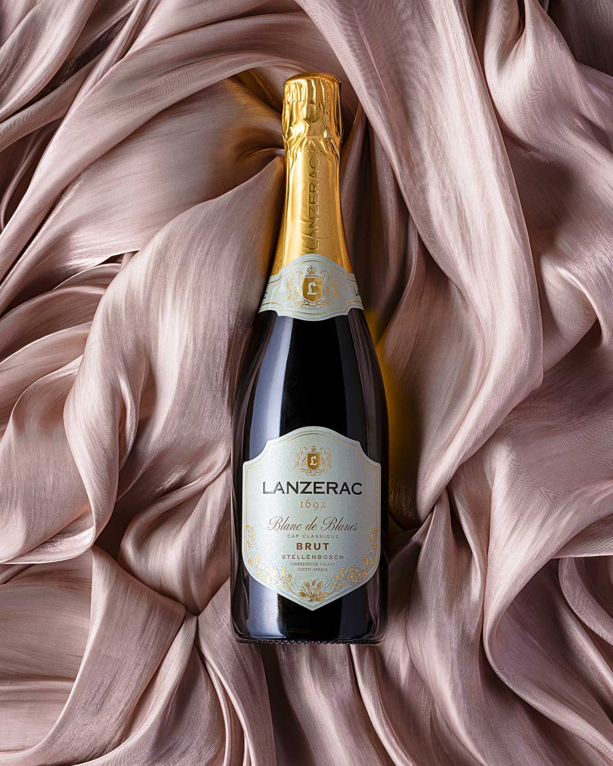Eine Flasche des Schaumweins Lanzerac 1692 Blanc de Blancs Brut ruht auf einem sanft drapierten, blassrosa Satinstoff.
