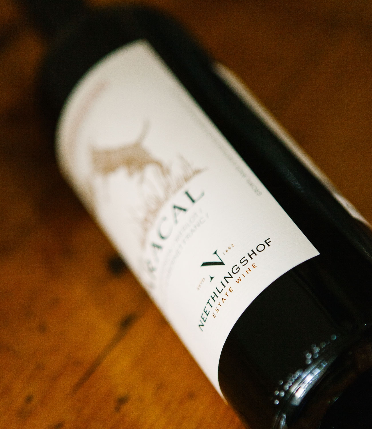 Eine Rotweinflasche mit der Aufschrift Der Karakal aus Neethlingshofs Kurzgeschichtensammlung, auf der ein Karakal abgebildet ist, der sich auf einen Vogel stürzt. Die Flasche hat einen schwarzen Verschluss und ein schlichtes, elegantes Design.