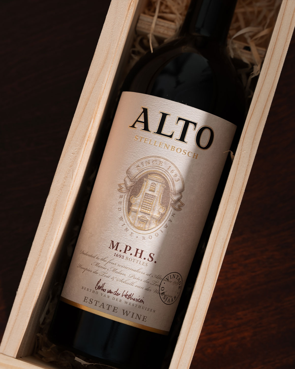 Eine Flasche Alto Stellenbosch Rotwein mit einem cremefarbenen Etikett mit goldener und weinroter Schrift, einem Wappen und der Aufschrift Estate Wine am unteren Rand. Die Flasche ist mit einer kastanienbraunen Folie verschlossen.