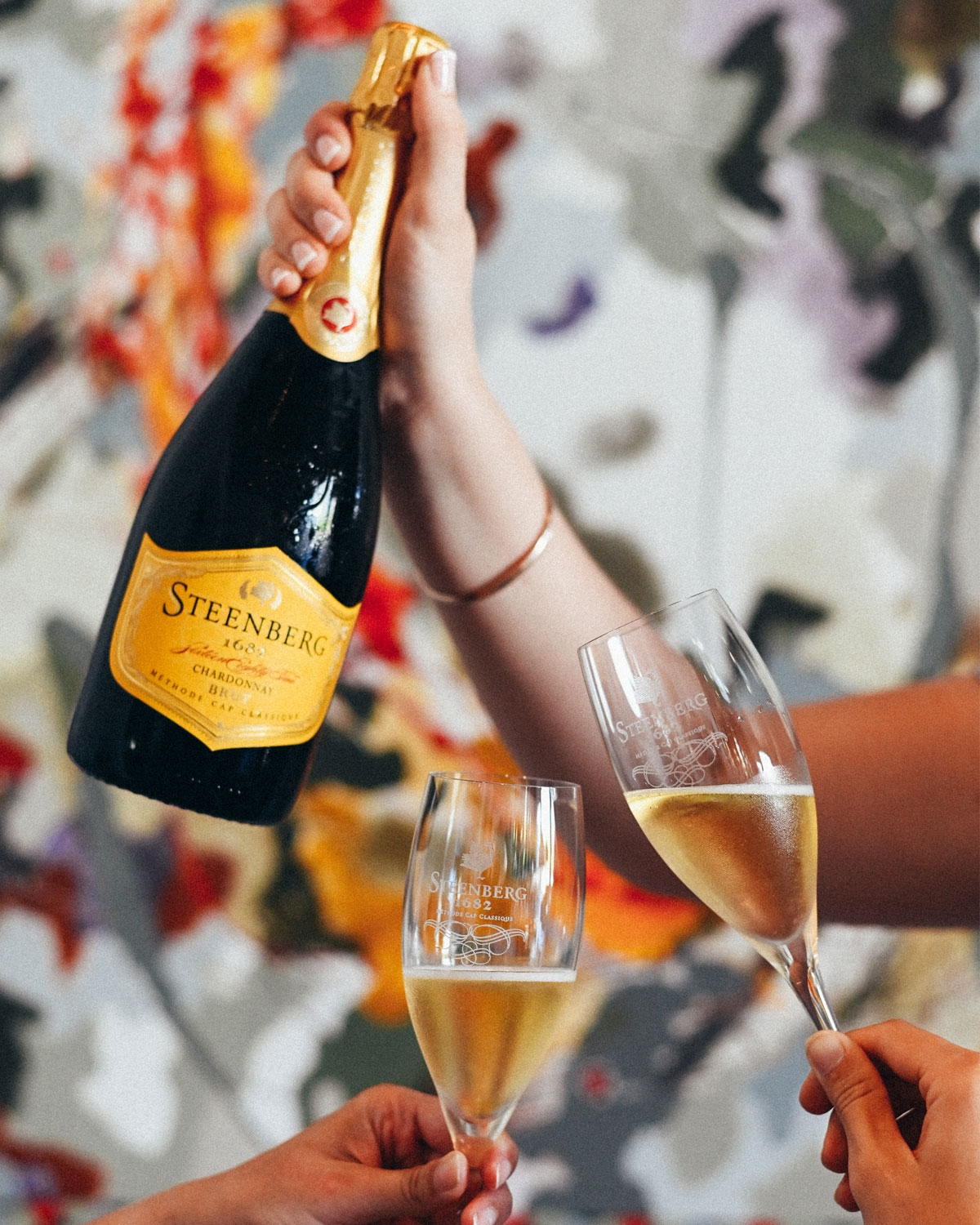 Eine Hand hält eine Flasche Steenberg-Sekt, während zwei andere Hände vor einem unscharfen, farbenfrohen Hintergrund auf gefüllte Weingläser anstoßen.