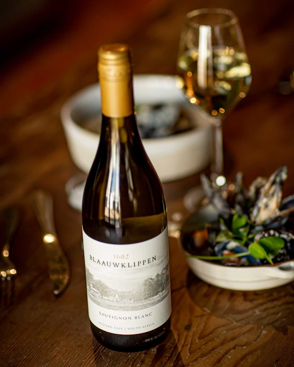 Eine Flasche Blaauwklippen 1682 Sauvignon Blanc aus dem Westkap, Südafrika, mit einem beigen Etikett, auf dem ein Weinberg mit Bergen im Hintergrund abgebildet ist.