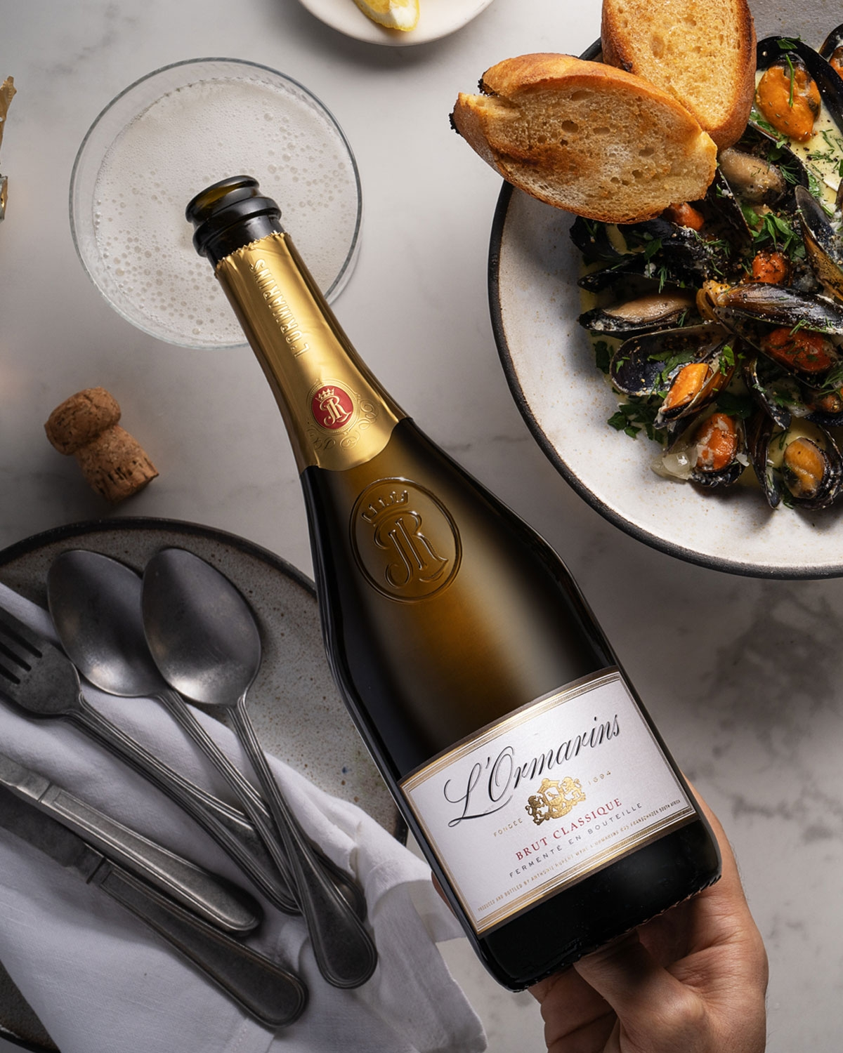 Eine Hand hält eine Flasche LOrmarins Brut Classique über einem Tisch, auf dem ein Cocktail, geröstetes Brot, eine Schale mit Muscheln, ein Teller mit Löffeln und eine Serviette stehen.