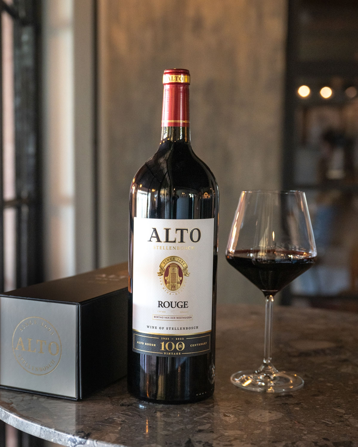 Eine Flasche Alto Stellenbosch Rouge mit einem schwarz-goldenen Etikett, auf dem ein goldenes Emblem und der 100. Jahrgang dieses südafrikanischen Weins gefeiert wird.