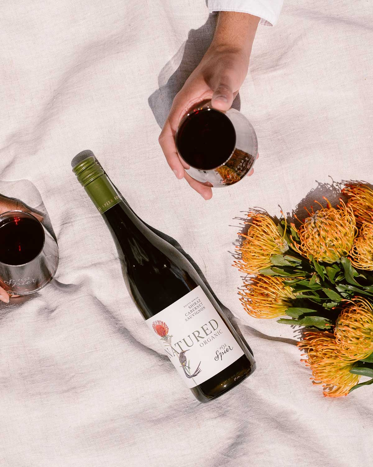 Eine Flasche Spier Natured Bio-Rotwein mit einem botanischen Etikett, das eine rote Blume und einen Vogel zeigt. Oben rechts befindet sich ein grünes Zertifizierungsetikett mit der Aufschrift WINE FROM NON-EU AGRICULTURE auf Englisch und Deutsch.