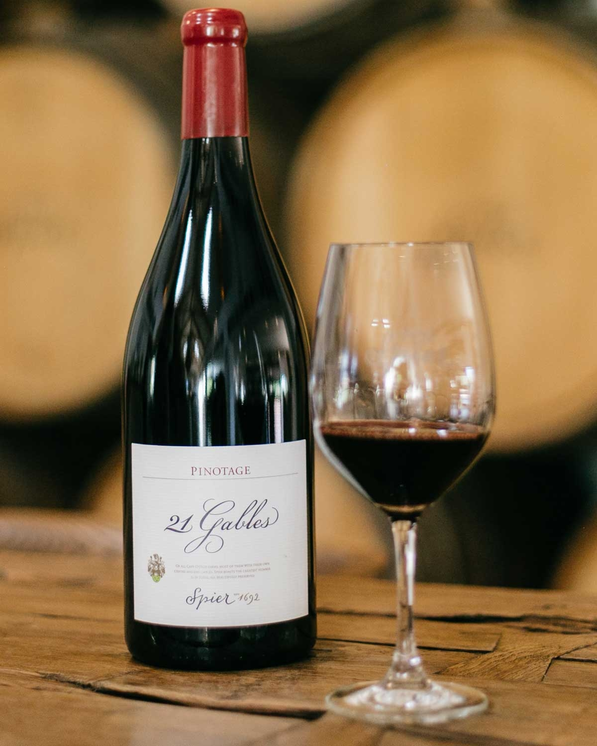 Eine Flasche 21 Gables Pinotage-Wein mit rotem Verschluss steht auf einem Holztisch neben einem Weinglas, das mit Rotwein gefüllt ist. Große Holzfässer sind unscharf im Hintergrund zu erkennen.