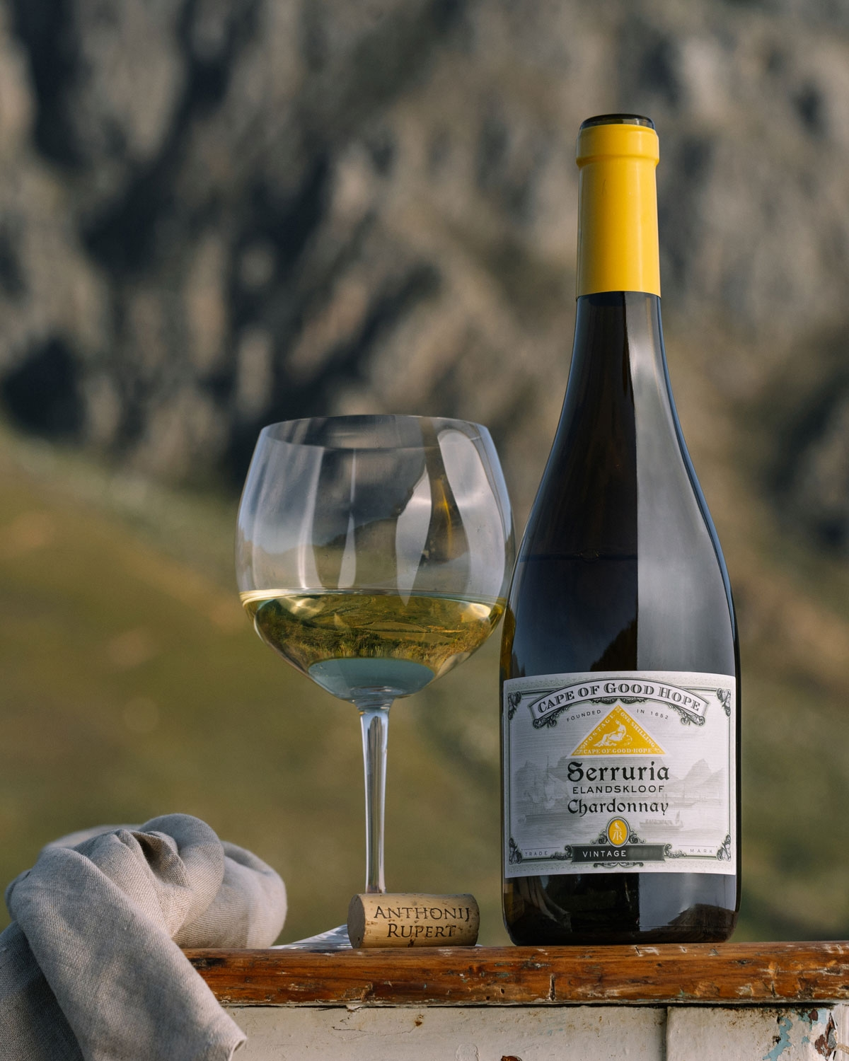 Eine Flasche Serruria Chardonnay-Wein und ein gefülltes Glas stehen auf einer rustikalen Fläche mit Korken und grauem Tuch vor einem unscharfen, bergigen Hintergrund im Freien.
