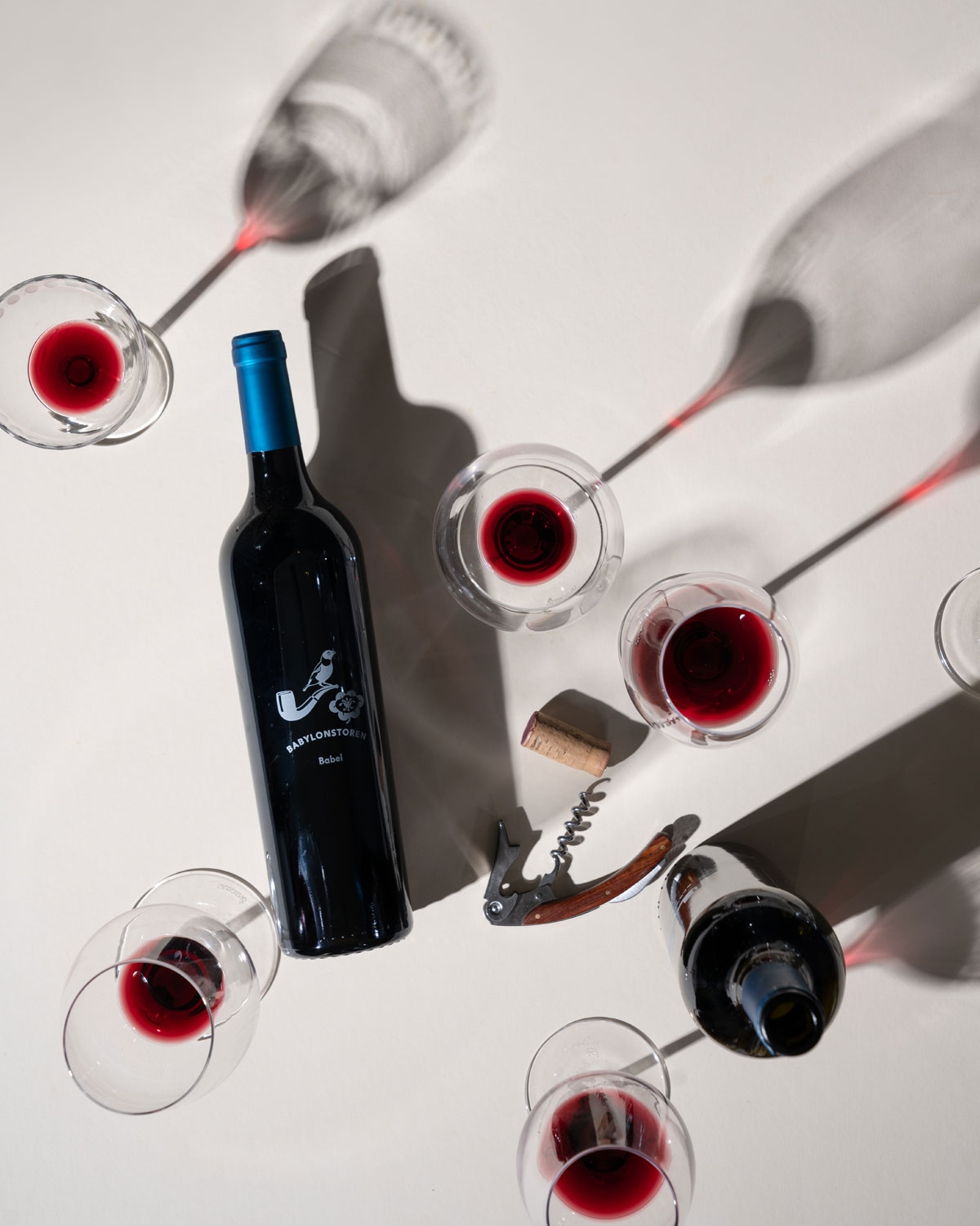 Eine Rotweinflasche, umgeben von fünf teilweise mit Rotwein gefüllten Gläsern, einem Korken und einem Korkenzieher auf einer hellen Fläche, die Schatten wirft.