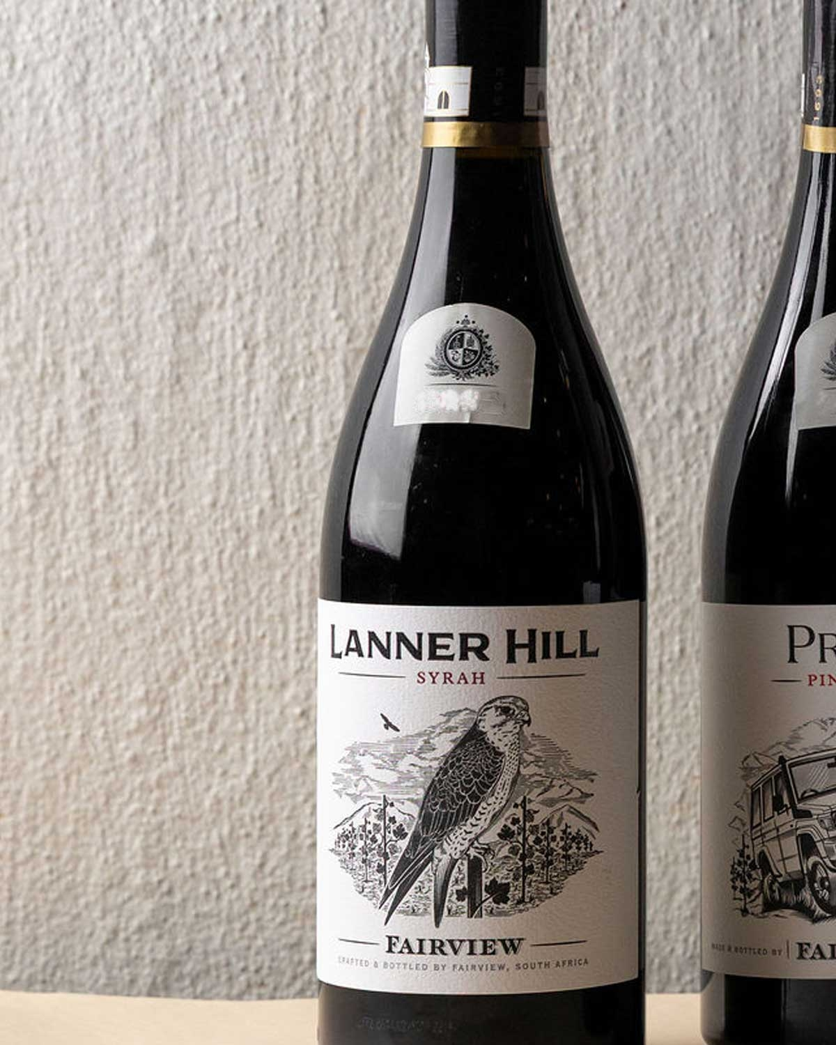 Eine Flasche Lanner Hill Syrah aus Fairview, Südafrika, mit einem Etikett, auf dem ein Vogel auf einem Zweig sitzt, vor einem strukturierten, cremefarbenen Hintergrund.