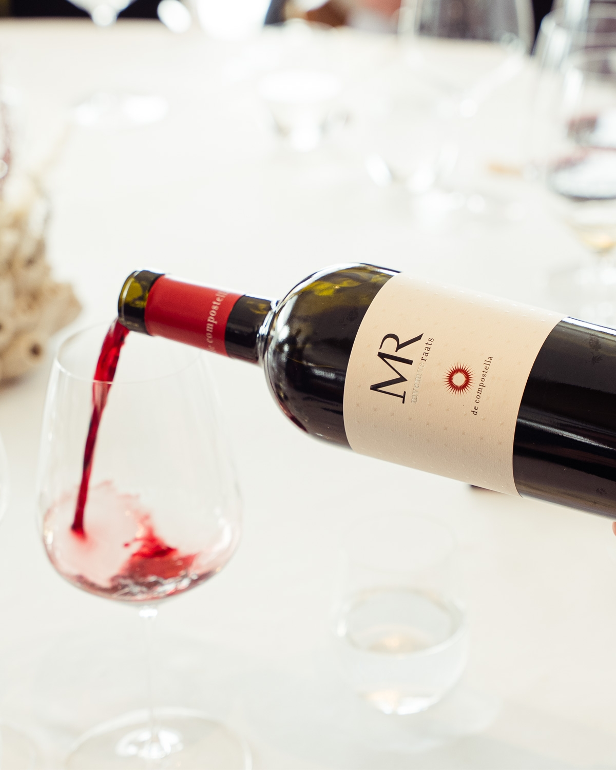 Eine Hand gießt Rotwein aus einer Flasche mit der Aufschrift MR in ein klares Weinglas auf einem weißen Tischtuch, mit Wassergläsern und unscharfen Tischgegenständen im Hintergrund.