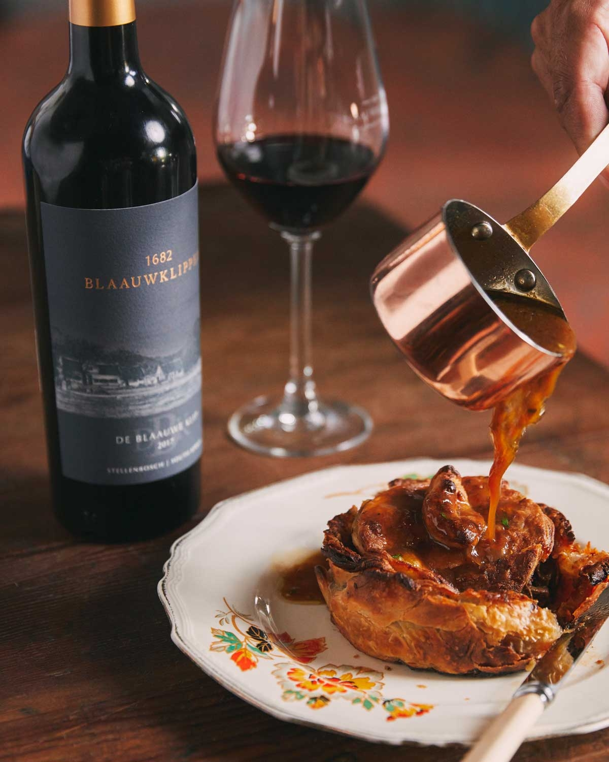 Eine Flasche Blaauwklippen-Rotwein mit einem Goldfolienverschluss und einem dunklen Etikett mit der Aufschrift 1682 Blaauwklippen und einer Landschaftsillustration, aus Stellenbosch, Südafrika.