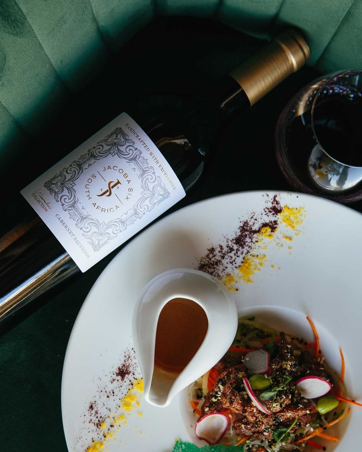Eine Flasche südafrikanischer Jacobs-Bay-Wein steht neben einem Glas Rotwein und einem Gourmetsalat mit Radieschen, Grünzeug und einer kleinen weißen Sauciere auf einem weißen Teller, garniert mit bunten Pülverchen.