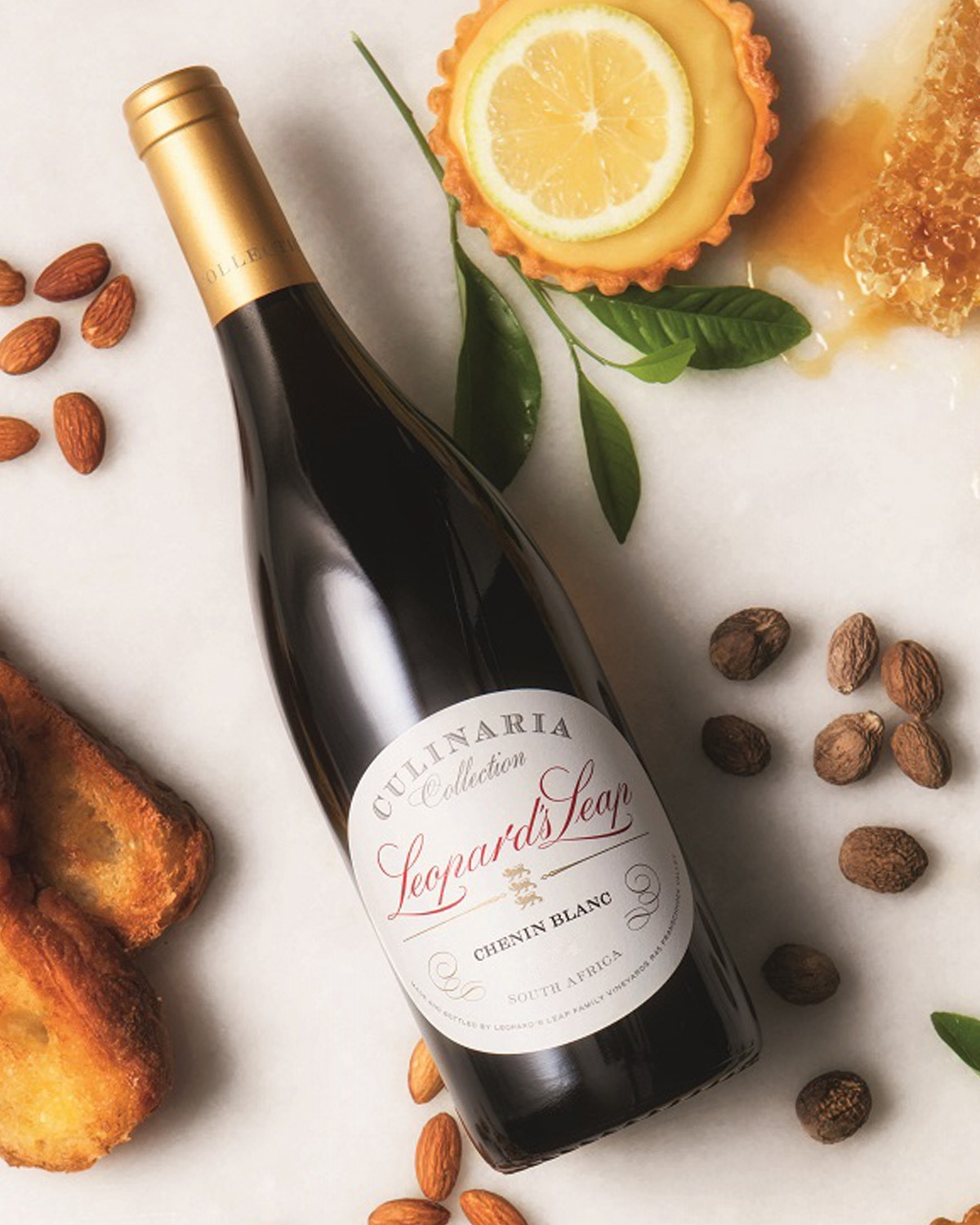 Eine Flasche Chenin Blanc der Leopard's Leap Culinaria Collection liegt auf einer Marmorplatte, umgeben von Mandeln, Muskatnuss, Toast, Zitronenkuchen, Zitronenscheiben, grünen Blättern und Honigwaben.