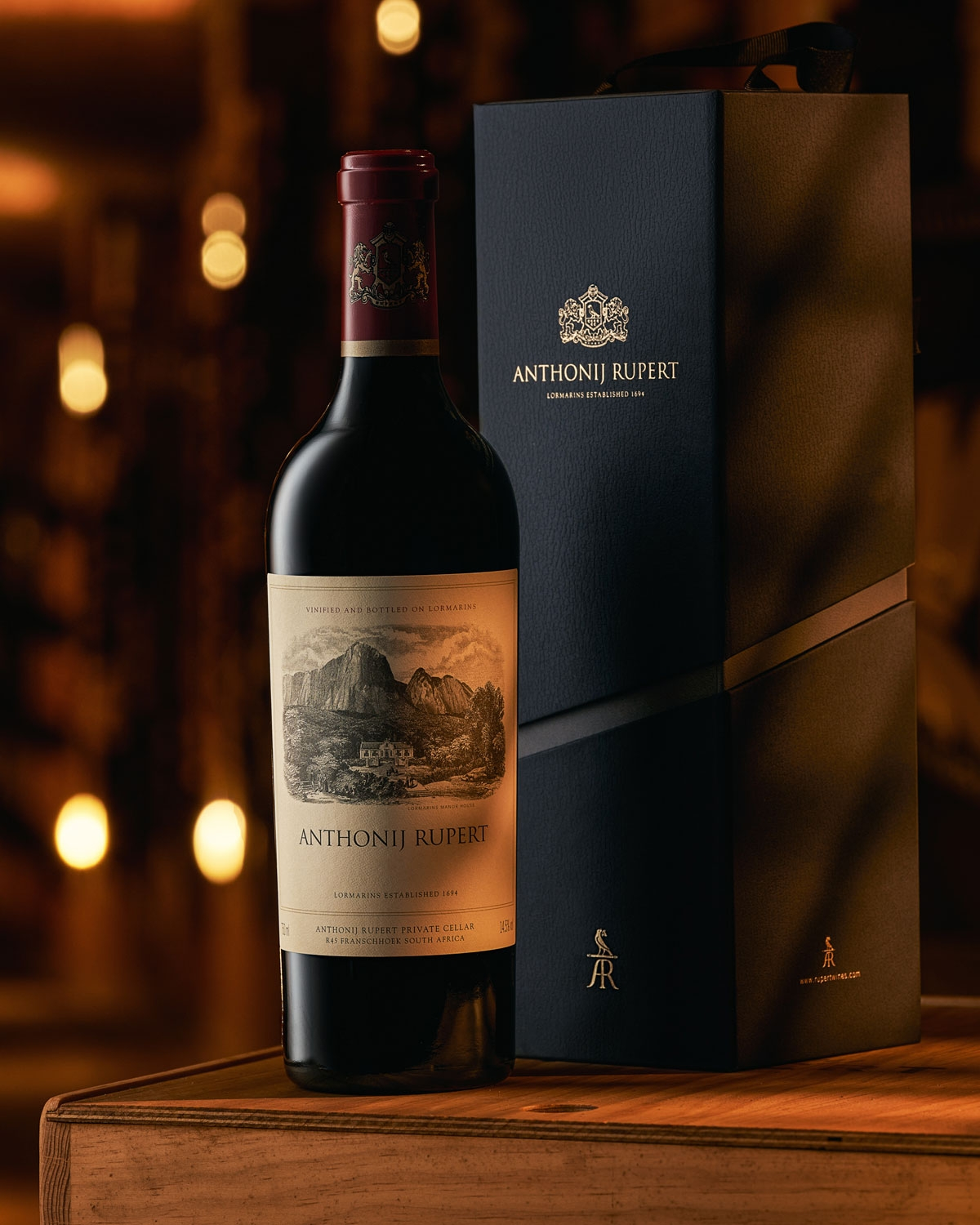 Eine Flasche Rotwein von Anthonij Rupert steht neben ihrer eleganten schwarzen Geschenkverpackung. Warme, sanft verschwommene Lichter im Hintergrund schaffen eine einladende, luxuriöse Atmosphäre.
