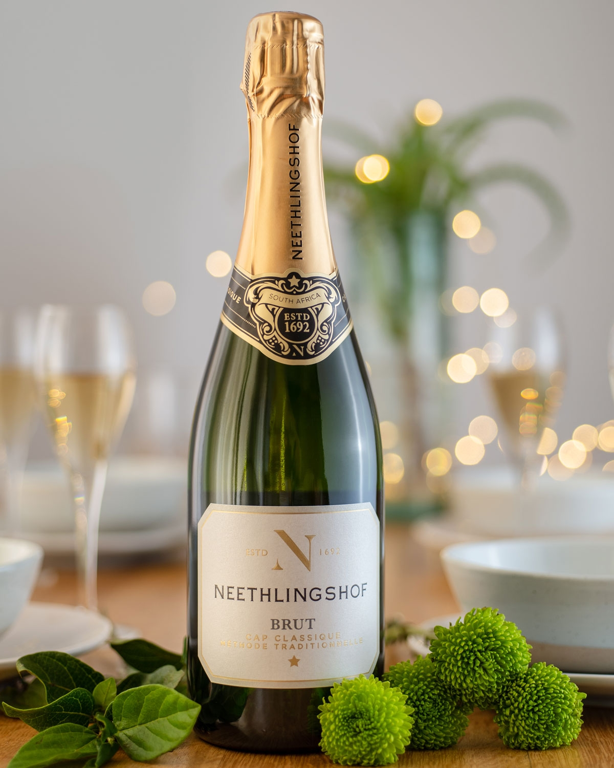 Eine Flasche Neethlingshof Brut Sekt steht auf einem Tisch mit grünen Blumen, Blattgarnituren, zwei gefüllten Sektgläsern, weißen Schalen und sanften Lichterketten im Hintergrund.