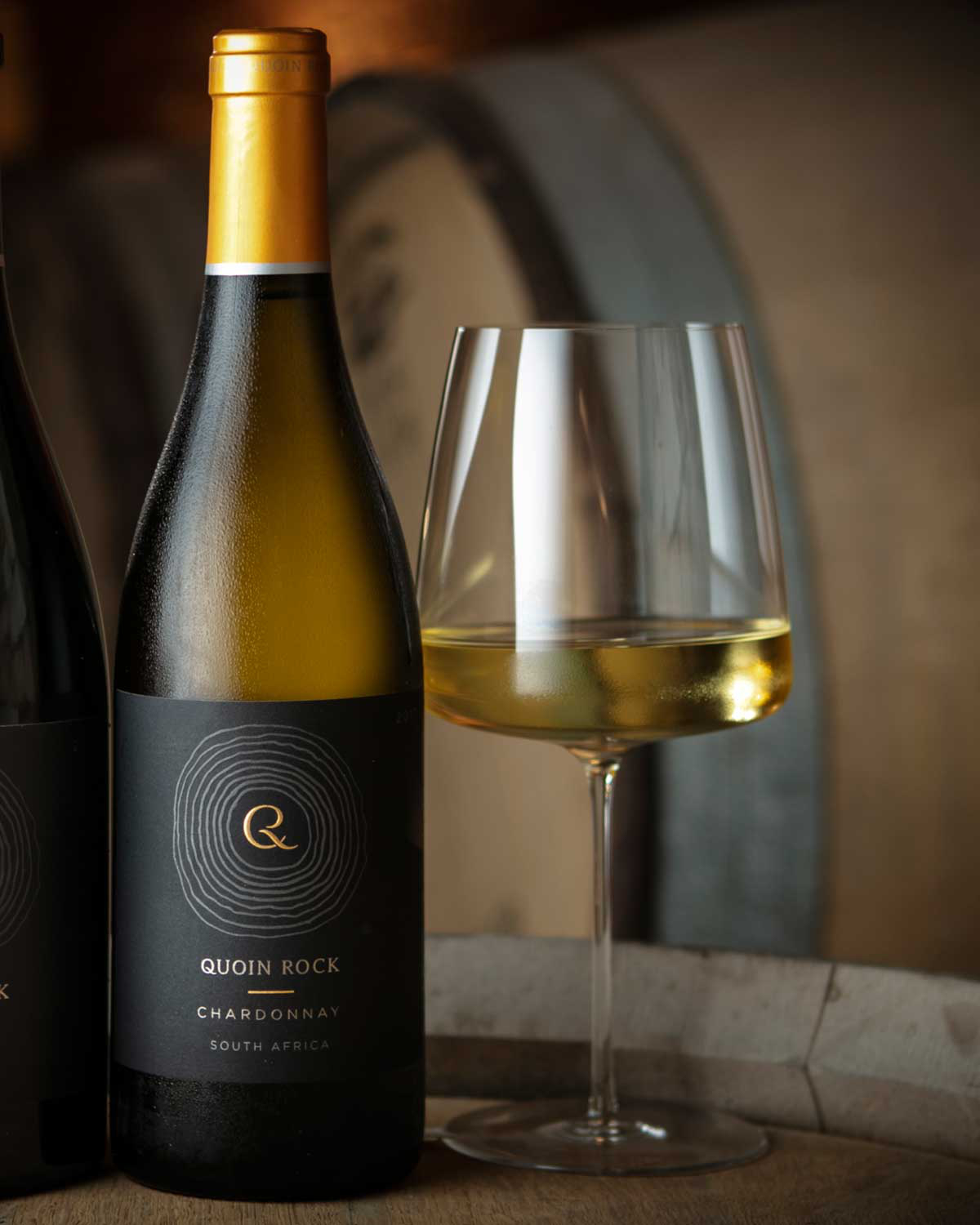 Eine Flasche Quoin Rock Chardonnay aus Südafrika mit einem goldenen Verschluss und einem schwarzen Etikett mit kreisförmigem Muster und dem Markennamen in weißer und goldener Schrift.