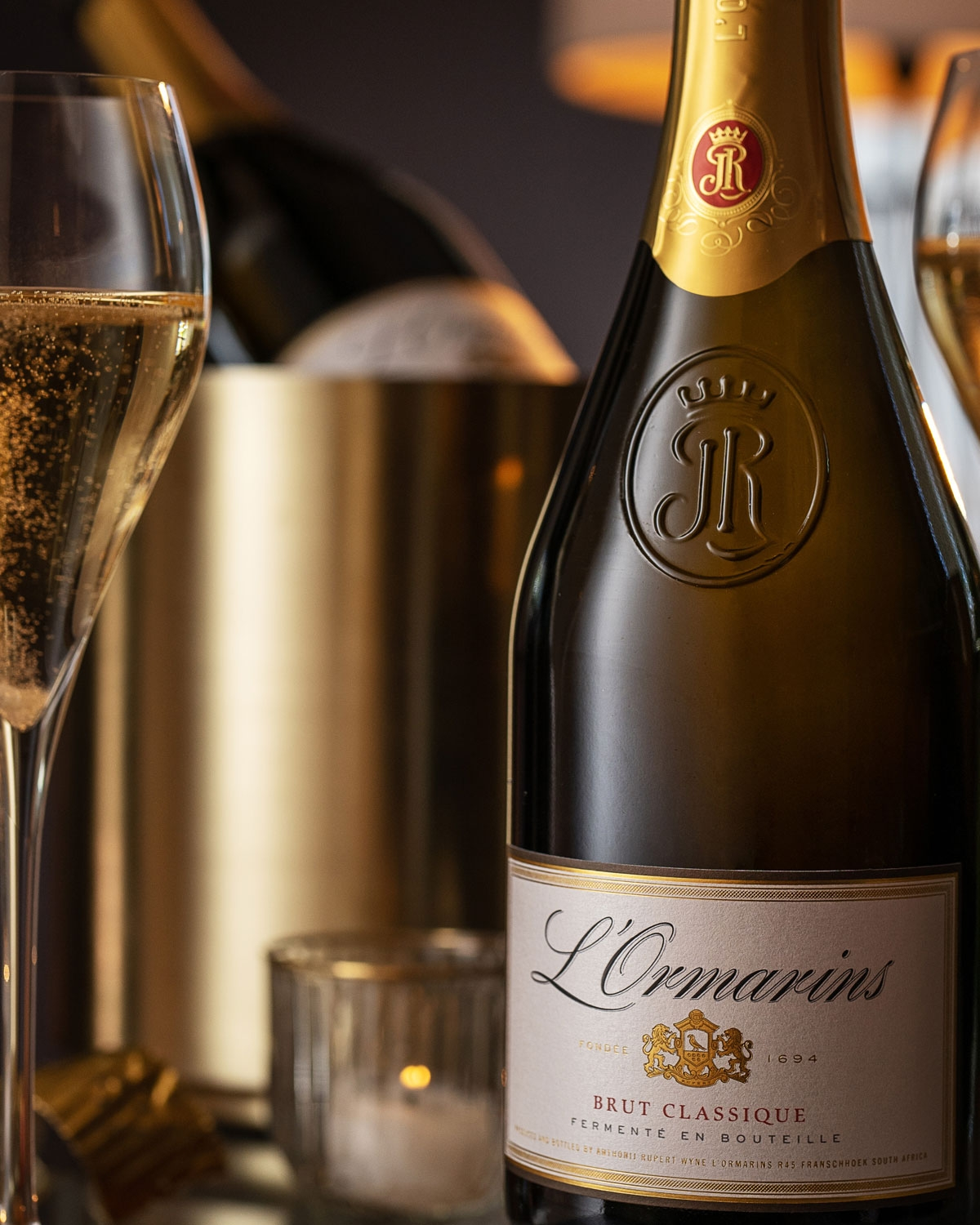 Eine Nahaufnahme einer L'Ormarins Brut Classique Sektflasche, eine gefüllte Champagnerflöte und ein goldener Eiskübel mit weiteren Flaschen im Hintergrund, die alle auf einem eleganten Tisch stehen.