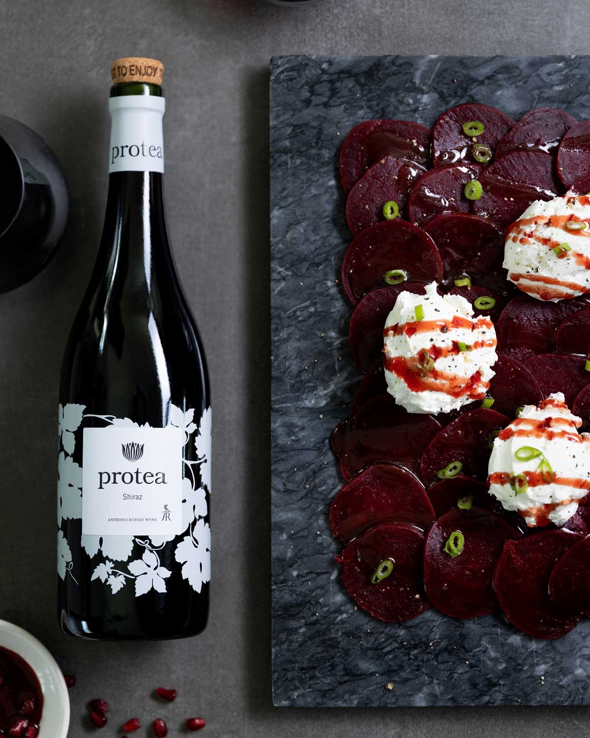 Eine Flasche Protea Shiraz-Wein steht neben einer Schieferplatte mit geschnittenen Rüben, Käsescheiben, grünen Zwiebeln und einem Spritzer Soße. Ein Glas Rotwein und eine kleine Schale sind teilweise sichtbar.