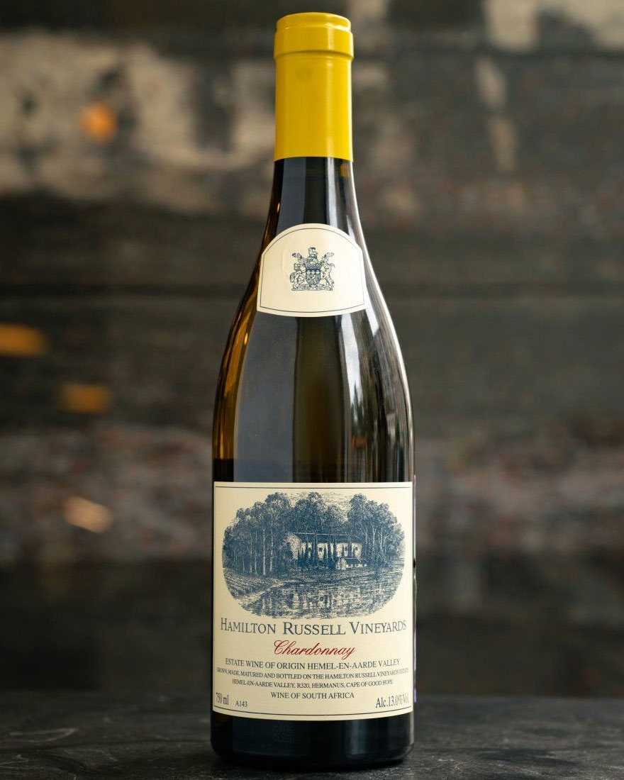 Eine Flasche Hamilton Russell Vineyards Chardonnay Wein mit einem cremefarbenen und blauen Etikett, auf dem ein Weinberg abgebildet ist und ein Text darauf hinweist, dass es sich um einen südafrikanischen Gutswein aus dem Hemel-en-Aarde Valley handelt.