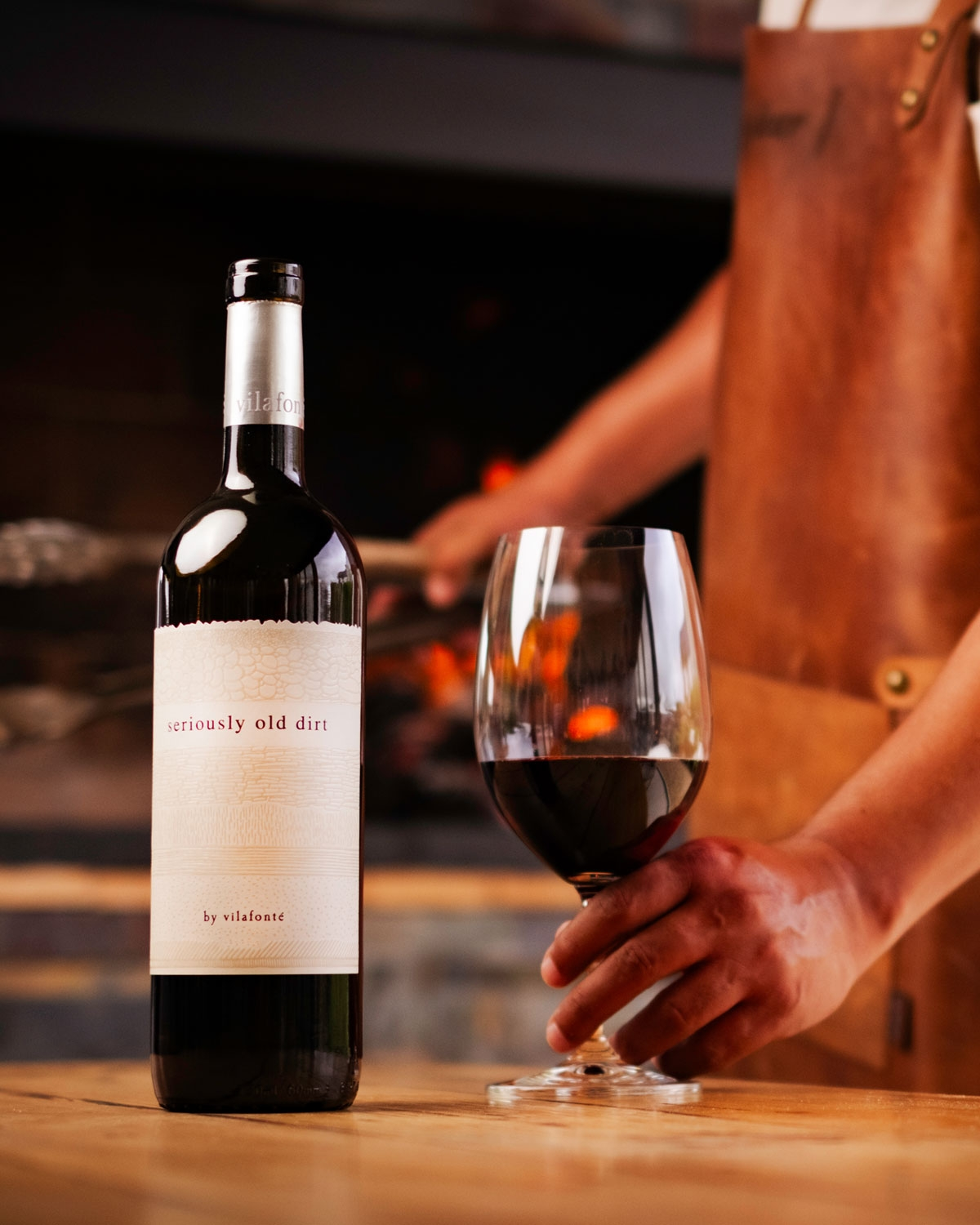 Eine Person in einer braunen Schürze hält ein Glas Rotwein neben einer Flasche mit der Aufschrift "seriously old dirt" von villafonté, mit einem warmen, verschwommenen Hintergrund.