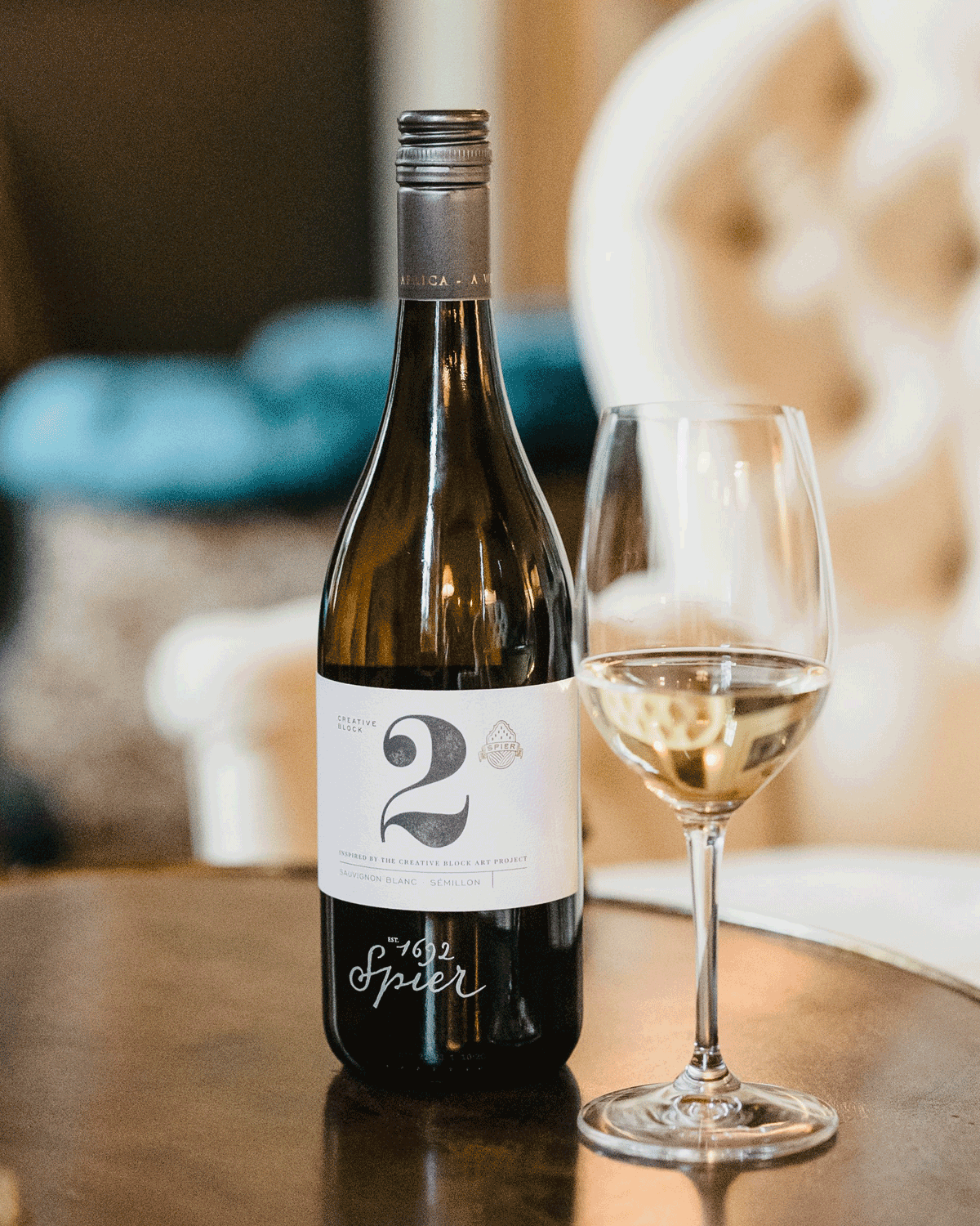 Eine Weißweinflasche mit der Aufschrift Spier 2021 steht neben einem gefüllten Weinglas auf einem Holztisch, mit einem unscharfen Plüschsessel und einem blauen Sofa im Hintergrund.