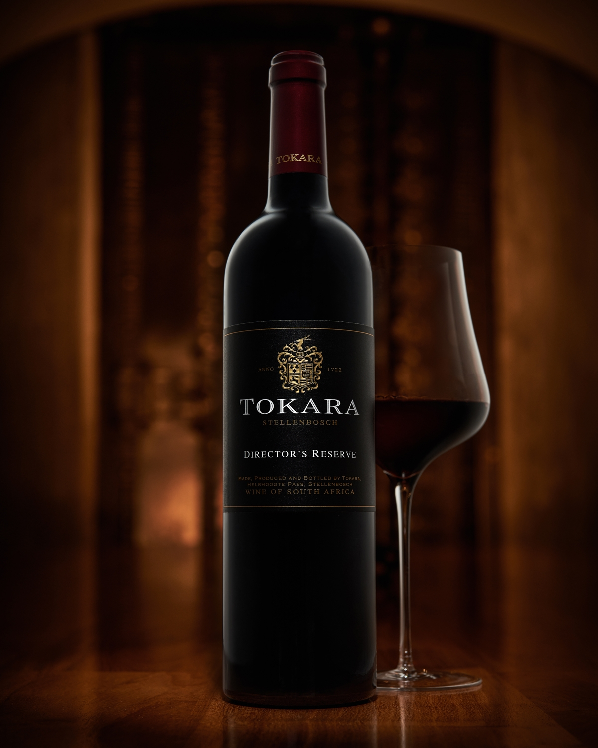 Eine Flasche Tokara Director's Reserve Rotwein steht neben einem gefüllten Weinglas auf einer hölzernen Oberfläche. Ein warmer, sanft beleuchteter Hintergrund schafft eine elegante Atmosphäre.