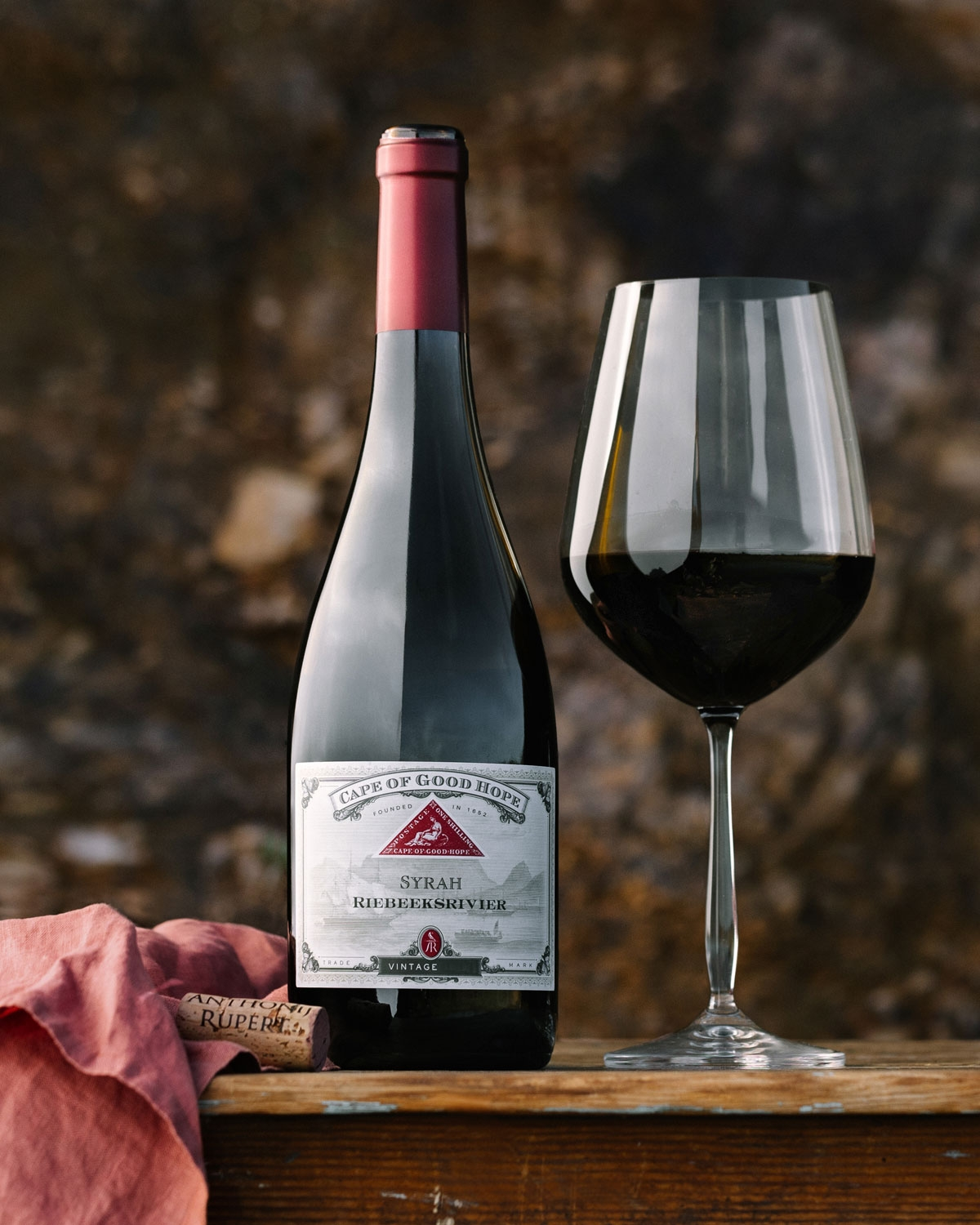 Eine Flasche Syrah-Wein vom Kap der Guten Hoffnung steht neben einem gefüllten Weinglas auf einer hölzernen Oberfläche, mit einem Korken und einem roten Tuch in der Nähe vor einem verschwommenen braunen Hintergrund.