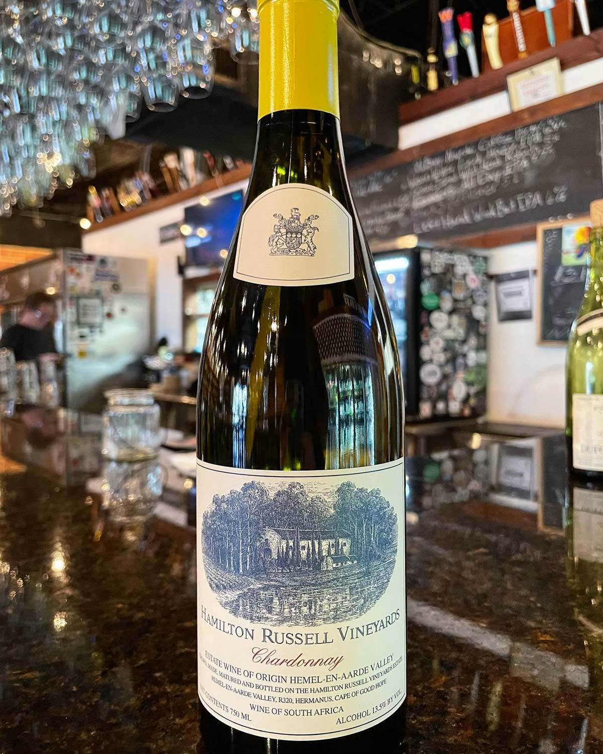 Eine Flasche Hamilton Russell Vineyards Chardonnay steht auf einem glänzenden Tresen in einem gemütlichen, schwach beleuchteten Restaurant, in dessen Hintergrund Regale, Weingläser und eine Speisekarte aus Kreide verschwimmen.