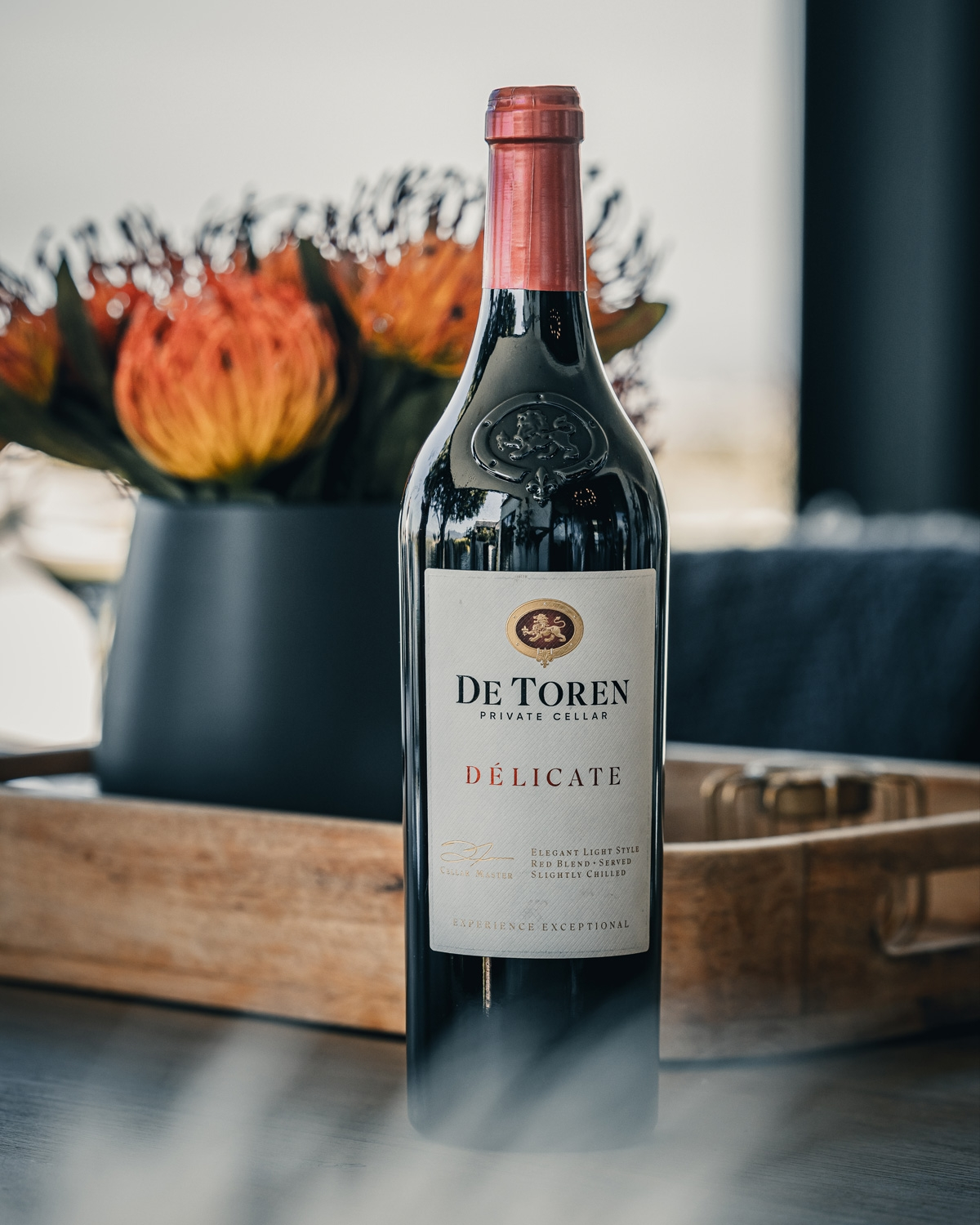 Eine Flasche De Toren Private Cellar Délicate Wein steht auf einem Tisch vor einem Holztablett und einer schwarzen Vase mit orangefarbenen Protea-Blüten. Das Etikett ist sichtbar und der Hintergrund ist leicht unscharf.