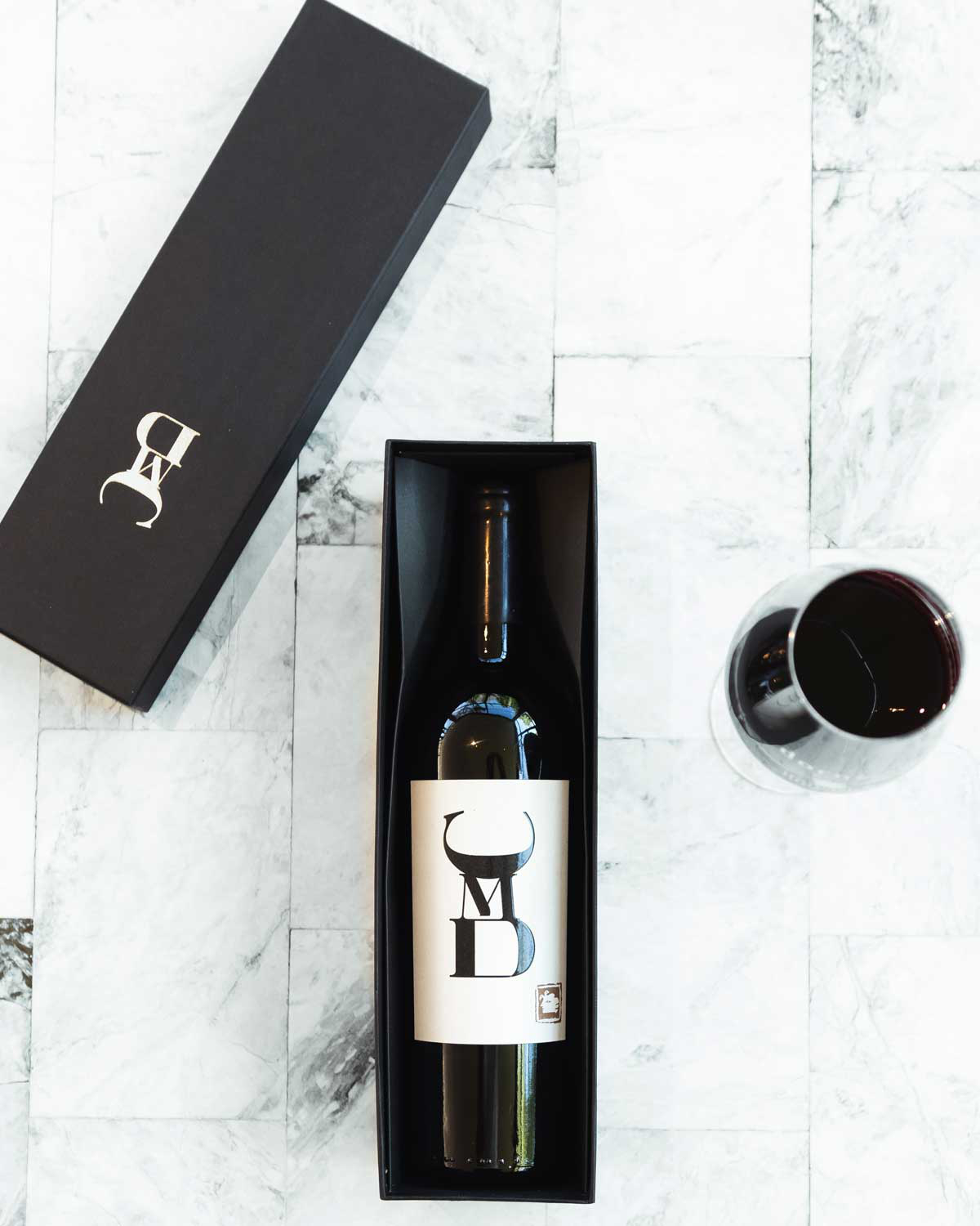 Eine Flasche Rotwein in einem schwarzen Geschenkkarton mit fetten schwarzen und weißen Buchstaben auf dem Etikett, platziert auf einer hellen Marmorfläche neben einem gefüllten Weinglas und dem Kartondeckel.