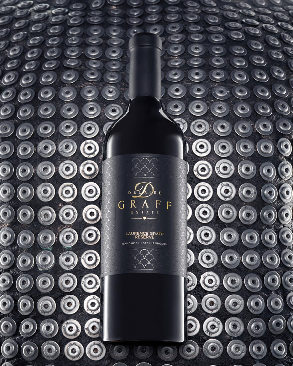 Eine Flasche Delaire Graff Estate Rotwein steht aufrecht vor einem Hintergrund aus metallischen, kreisförmigen Patronenhülsen, die in einem Gittermuster angeordnet sind. Das Weinetikett ist dunkel mit goldenen Akzenten.