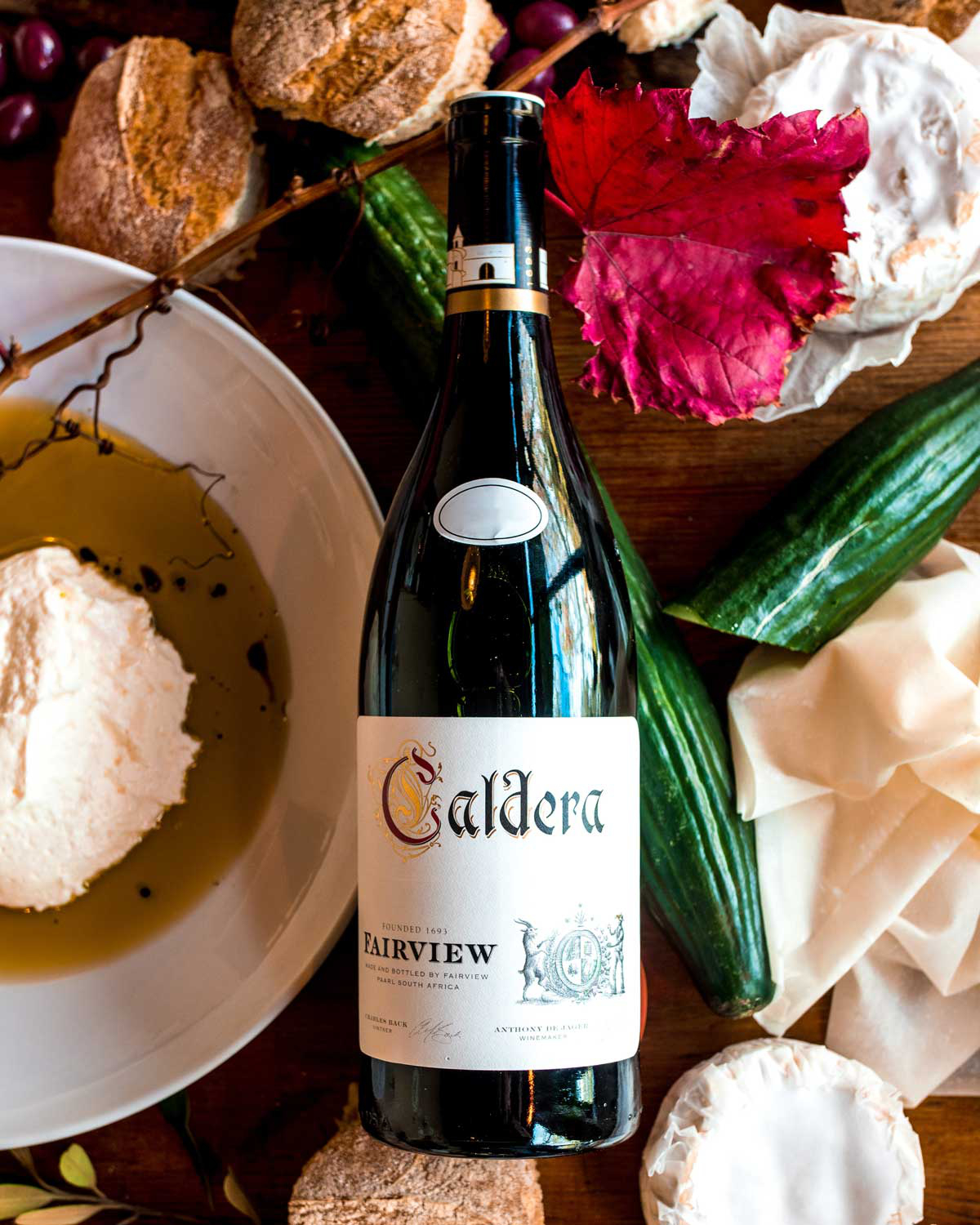 Eine Flasche Caldera Fairview Rotwein mit schwarzem Folienverschluss, goldenem und weißem Etikett und dekorativem Text, mit einer kleinen Illustration eines Gebäudes und einem Wappen am unteren Rand des Etiketts.